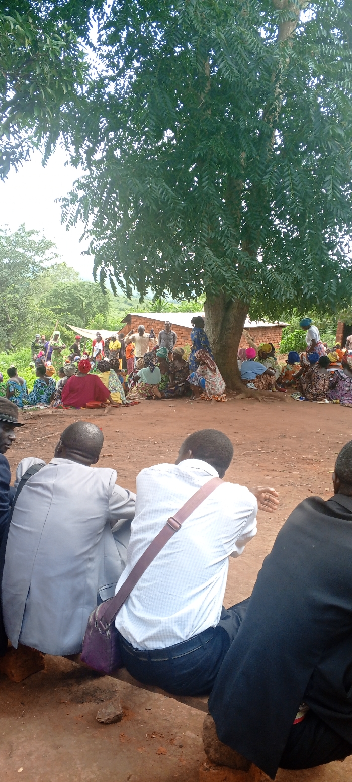 Under a large tree, a group of people are gathered in an outdoor setting. Most are seated on the ground, wearing colorful clothing, while others stand. The backdrop includes a building with a brick facade and surrounding green foliage. In the foreground, individuals dressed in formal attire, including suits and a shirt with a strap, are also seated, facing the larger group. The scene suggests a community meeting or gathering in a rural area.