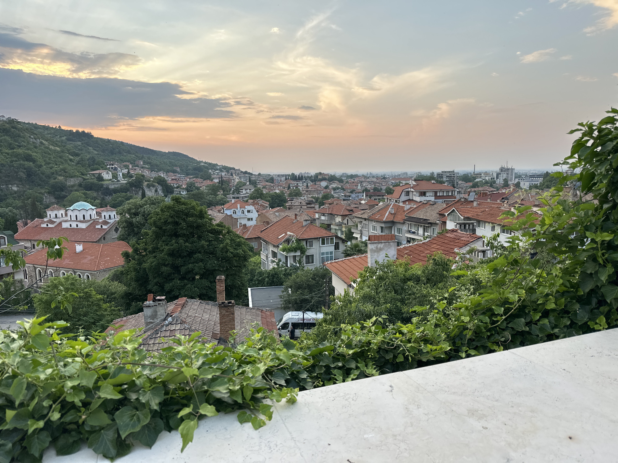 This elevated panoramic image captures a sprawling urban landscape in Asenovgrad, Bulgaria, observed from a vantage point, likely a balcony or terrace, during either sunrise or sunset. The sky displays a gradient of soft peach, orange, and light blue hues, with scattered stratocumulus clouds partially obscuring the sun, creating diffused light and subtle crepuscular rays.
In the immediate foreground, a white, textured stone railing or wall occupies the bottom right, partially covered by dense, vibrant green ivy with numerous leaves and tendrils extending upwards and horizontally.
The midground features a densely populated area of Asenovgrad. To the left, a steep, forested hill, part of the Rhodope Mountains, rises, covered in lush green deciduous trees. Houses with red-tiled roofs and light-colored facades are nestled along its lower slopes. A prominent architectural feature is a white Eastern Orthodox church with a distinctive light turquoise dome and several smaller white domes, surrounded by red-tiled roofs. Moving towards the center and right, the city extends across a varied topography. Numerous multi-story residential buildings, predominantly two to four stories, exhibit a uniform aesthetic with terracotta or red-orange tiled roofs, often featuring multiple chimneys, and light-toned plaster facades. Green trees and shrubs are interspersed throughout the urban fabric.
In one of the cropped sections (Crop 5), a white cargo van is partially visible, appearing to be parked or navigating a narrow street. The text "БГ КУРЬЕР ТР..." (likely "BG KURIER TRANSPORT") is partially legible on its side, further confirming the Bulgarian location. One person, dressed in dark clothing, is visible near the driver's side of the van.
The background reveals a hazy cityscape stretching into the distance, with the silhouettes of taller, more modern multi-story buildings and industrial structures, indicating the continued expansion of Asenovgrad into the more distant, flatter plains. The overall atmosphere is tranquil, characterized by the soft, ambient light of the early morning or late evening.