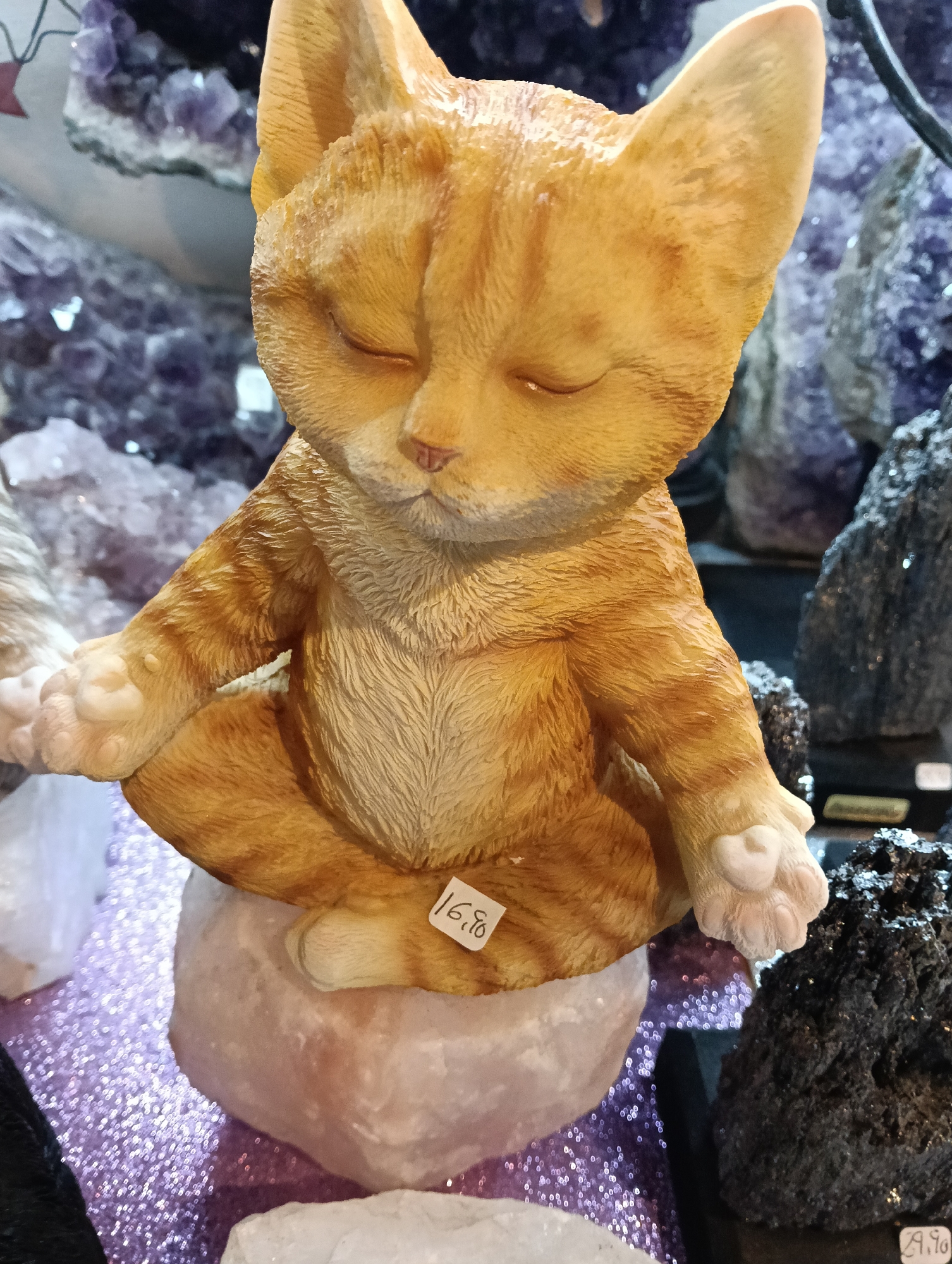 The image prominently features an orange tabby-like cat figurine, sculpted in a meditative pose with its eyes closed and front paws resting on its knees, palms facing upwards in a gesture often associated with mindfulness or prayer. The figurine exhibits carved fur texture and dark orange stripes, consistent with an tabby cat's markings. It is seated on a light pinkish-white, roughly hewn rock, possibly a salt lamp base or a large crystal.
In the background, various crystals and mineral specimens are visible. To the left and behind the cat, large formations of purple amethyst crystals can be seen, with their distinct crystalline structure. To the right and foreground of the background, darker, rough-textured rocks, possibly volcanic or metallic minerals, are displayed. The surface beneath the objects is a sparkly, glittery purple material.
A small white price tag is affixed to the cat figurine's left thigh, displaying the numbers "16,8" in black handwriting, likely indicating a price. Another small, illegible tag is partially visible on the dark rock in the bottom right corner. The overall scene suggests an indoor setting, likely a shop or market stall specializing in spiritual items, crystals, or gifts. The ambiance conveyed is one of tranquility and contemplation, primarily due to the meditating cat.