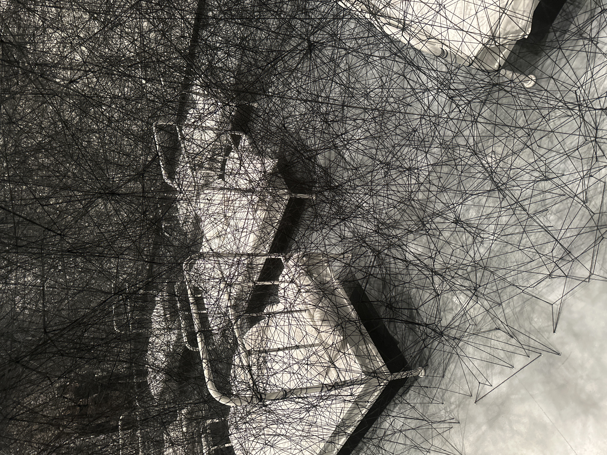 The image shows a close-up of a contemporary art installation. Thin black wires are stretched and connected to create a complex network of geometric shapes, partially obscuring a white metal structure resembling a bed or cot. The structure has rounded tubular elements. The background appears to be a gray surface, possibly the floor or a wall. The installation is located indoors, likely within an art gallery or museum. While the specific location in London is not discernible from the image, such installations are often found in art venues throughout the city.