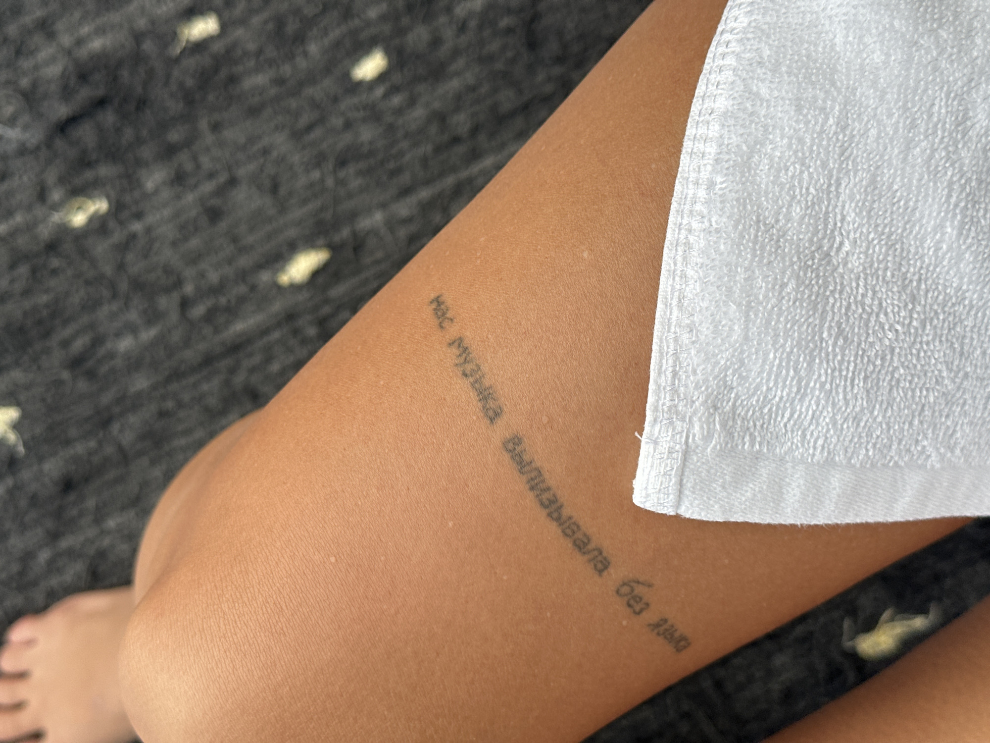 The image shows a person's thigh with a tattoo in Russian that reads "Нас музыка вылизывала без языка" ("Music licked us without a tongue"). A white towel partially covers the thigh. The person's foot is visible in the lower left corner of the image. The background is out of focus and appears to be dark gray with light yellow spots. There is no information about the location being Licin, Indonesia.