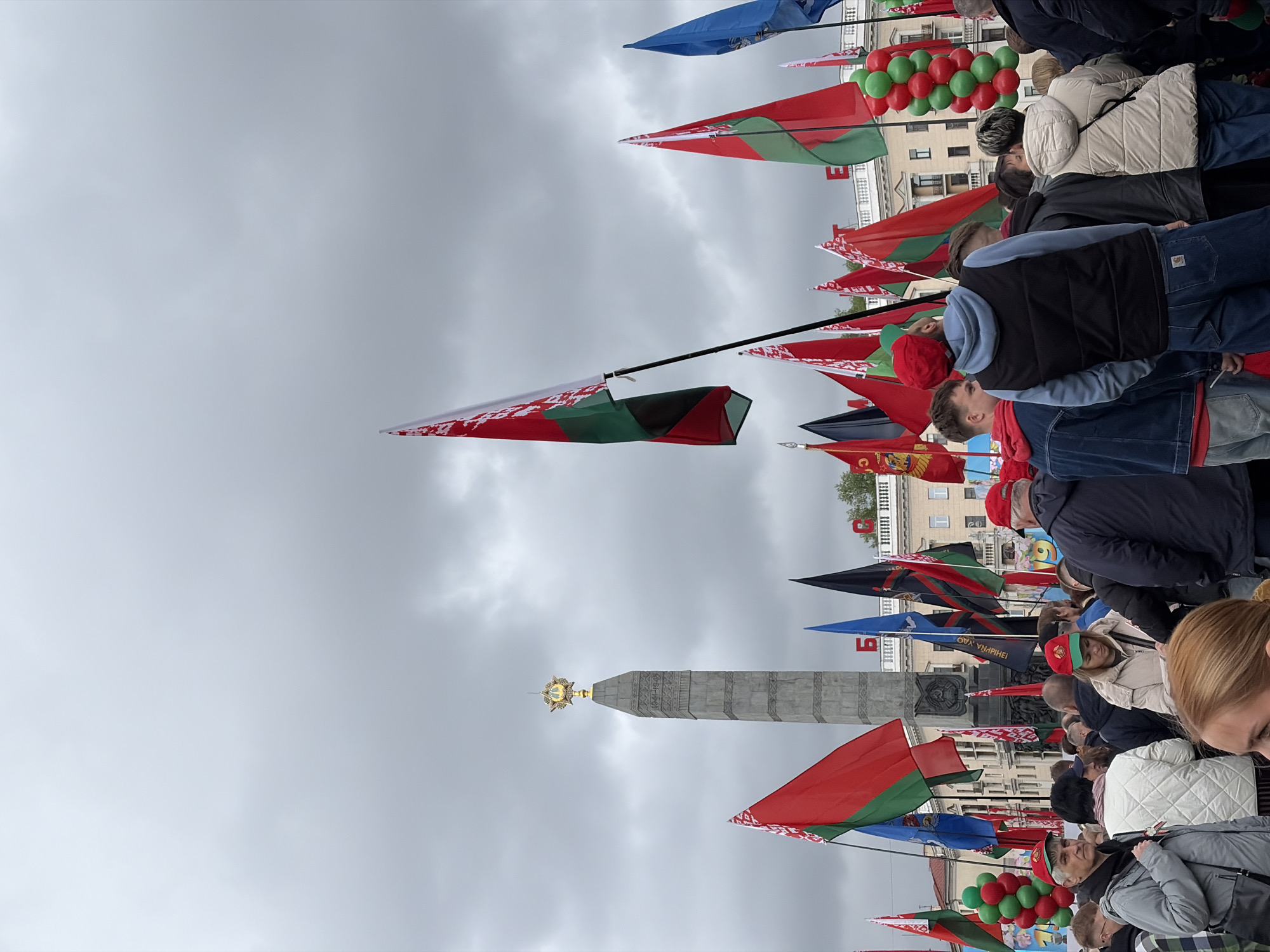 The image depicts a crowd of people gathered in an urban setting, possibly at a commemorative event or celebration. Several flags, primarily red and green, are visible, likely representing the Belarusian flag. A tall monument with a golden star on top stands prominently in the background, situated in front of a pale overcast sky. There are balloons in the colours red and green. The specific location can't be identified.