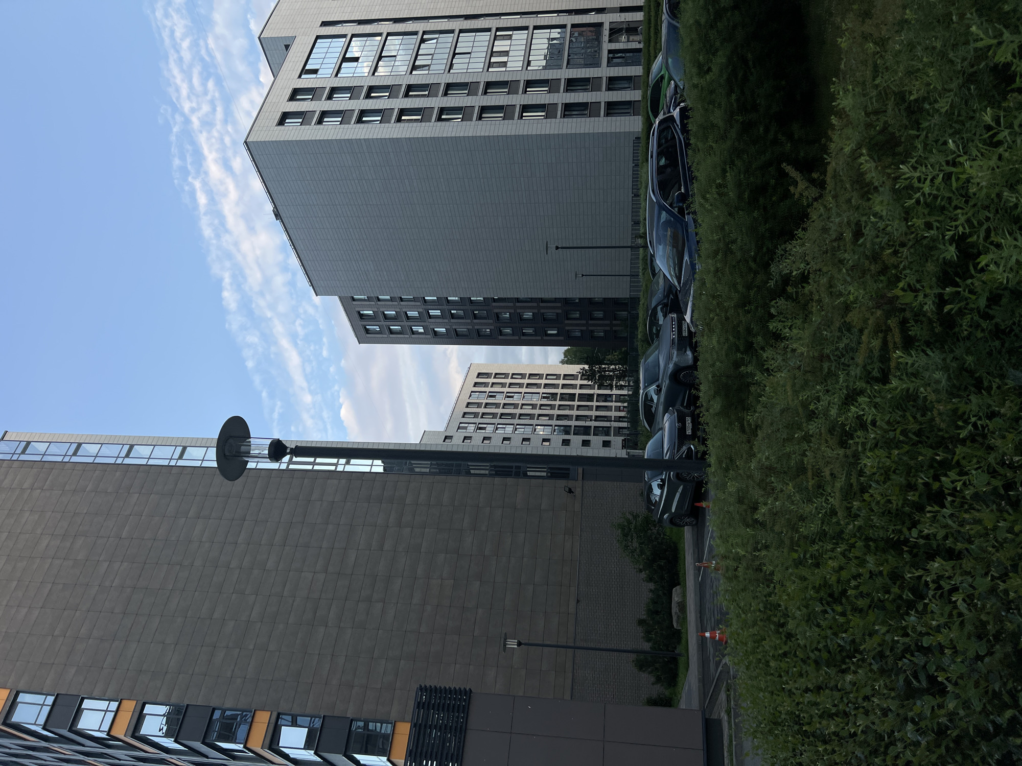 A low-angle, outdoor, full shot captures a modern residential complex in Saint Petersburg, Russia, under a partly cloudy blue sky. In the foreground, a dense, dark green hedge fills the bottom half of the frame. Beyond the hedge, a parking lot is visible with several cars parked. The cars are dark-colored SUVs and sedans. Orange traffic cones are placed on the asphalt. In the middle ground, two multi-story apartment buildings are prominently featured. The building on the left is taller and has a dark grey facade with a band of orange detailing near the top. It features numerous windows, some with blue reflections. A tall, modern lamppost stands in front of this building. To the right, a slightly shorter apartment building has a lighter grey facade and many windows that reflect the sky. Further in the background, another similar apartment building is visible, along with some trees. The overall scene depicts a contemporary urban environment with an emphasis on residential architecture.