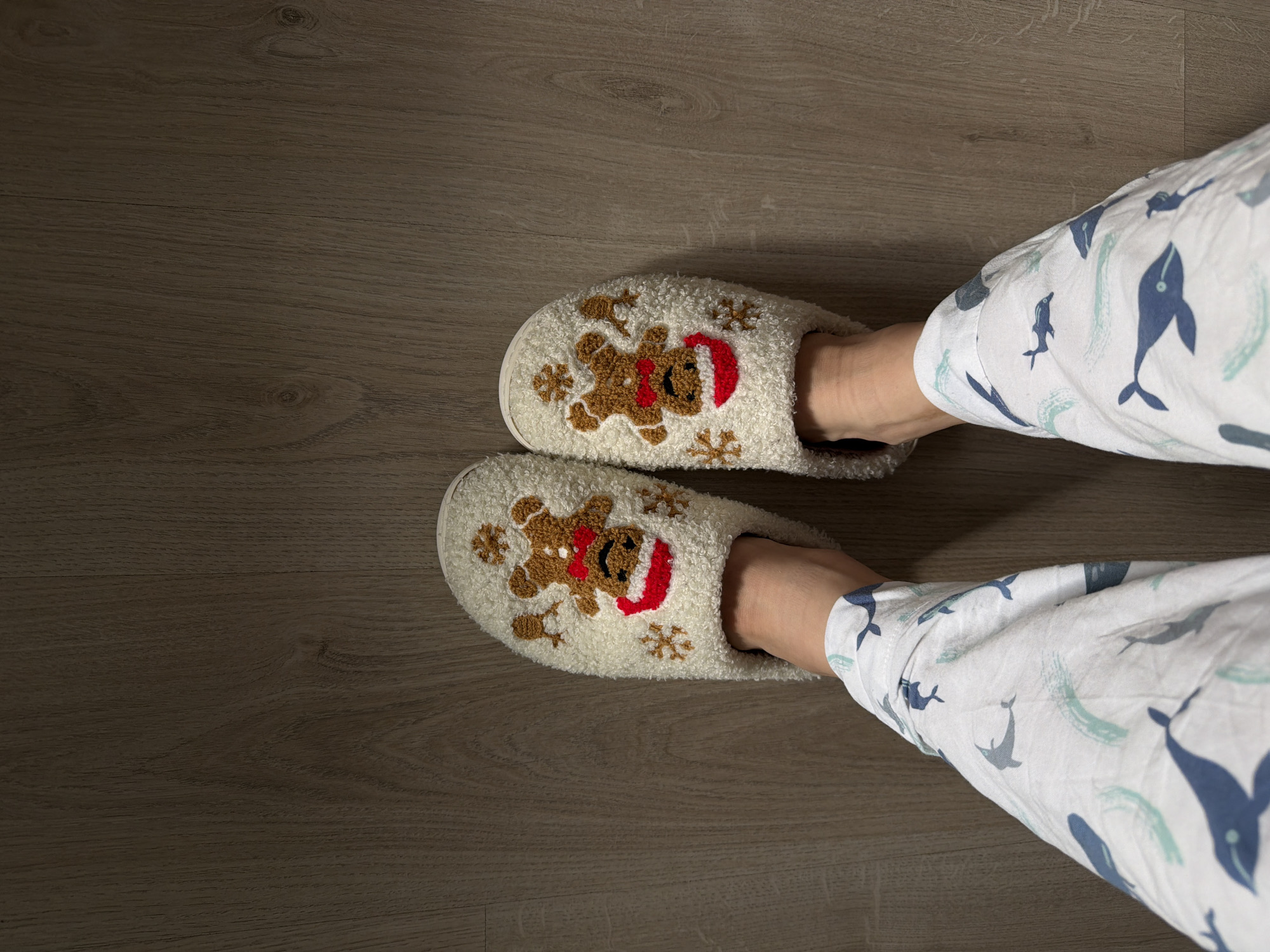 The image shows a person's feet in white, fuzzy slippers with gingerbread man designs. The person is wearing white pajamas with a whale pattern. They are standing on a wood-look floor. The location is unknown, and there is no information to indicate it is Izhevsk, Russia.