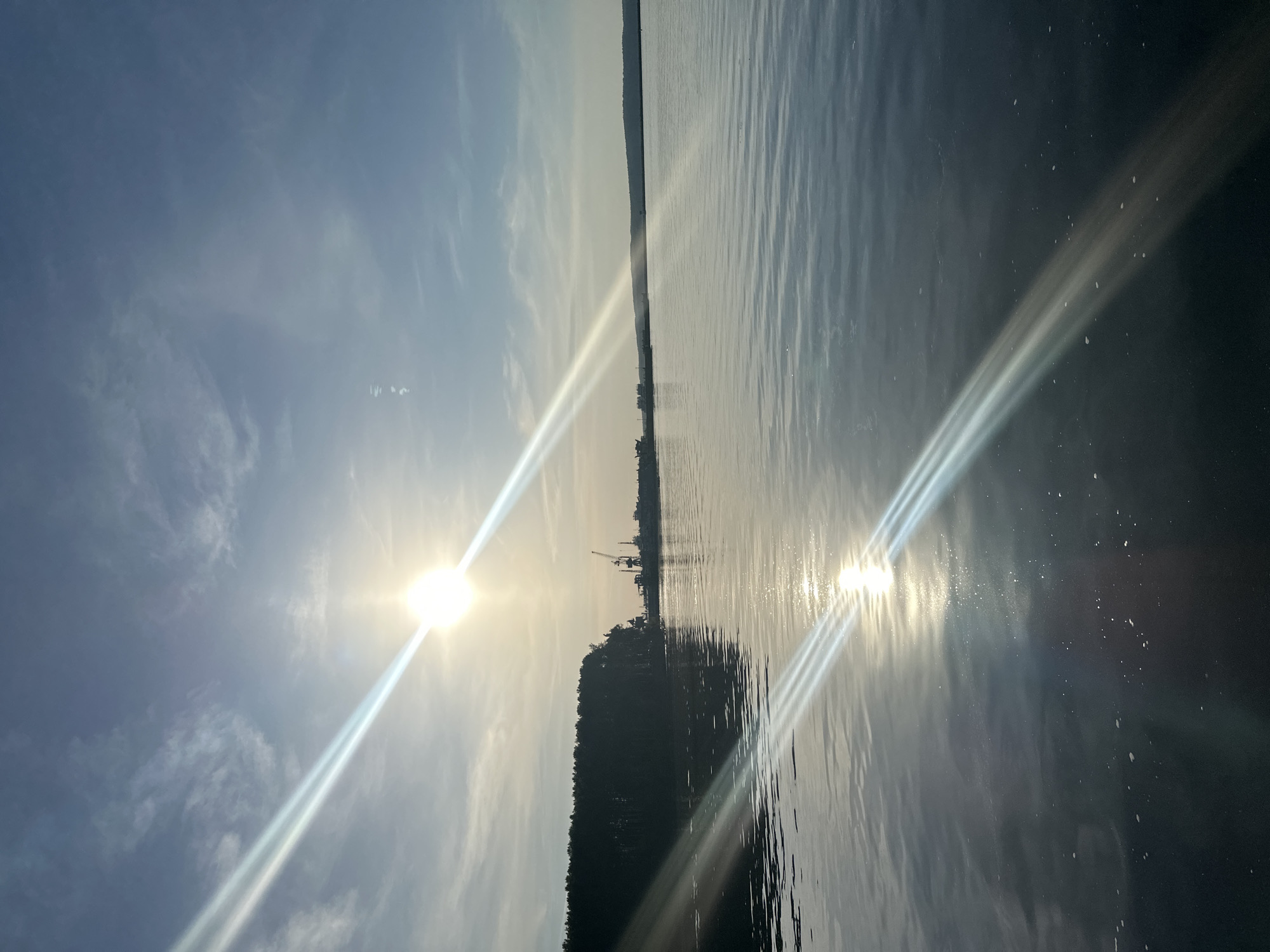 The image depicts a body of water, possibly a lake or a river, reflecting the sunlight. The sun is positioned in the sky, casting a bright glare with visible light rays. On the opposite shore, there appears to be a land mass covered with trees. In the distance, there are structures that resemble industrial cranes, suggesting a port or industrial area. The sky is partly cloudy with a pale blue hue. The water is slightly rippled, creating a textured surface that enhances the reflection of the sun. The location is identified as Unknown, Unknown.