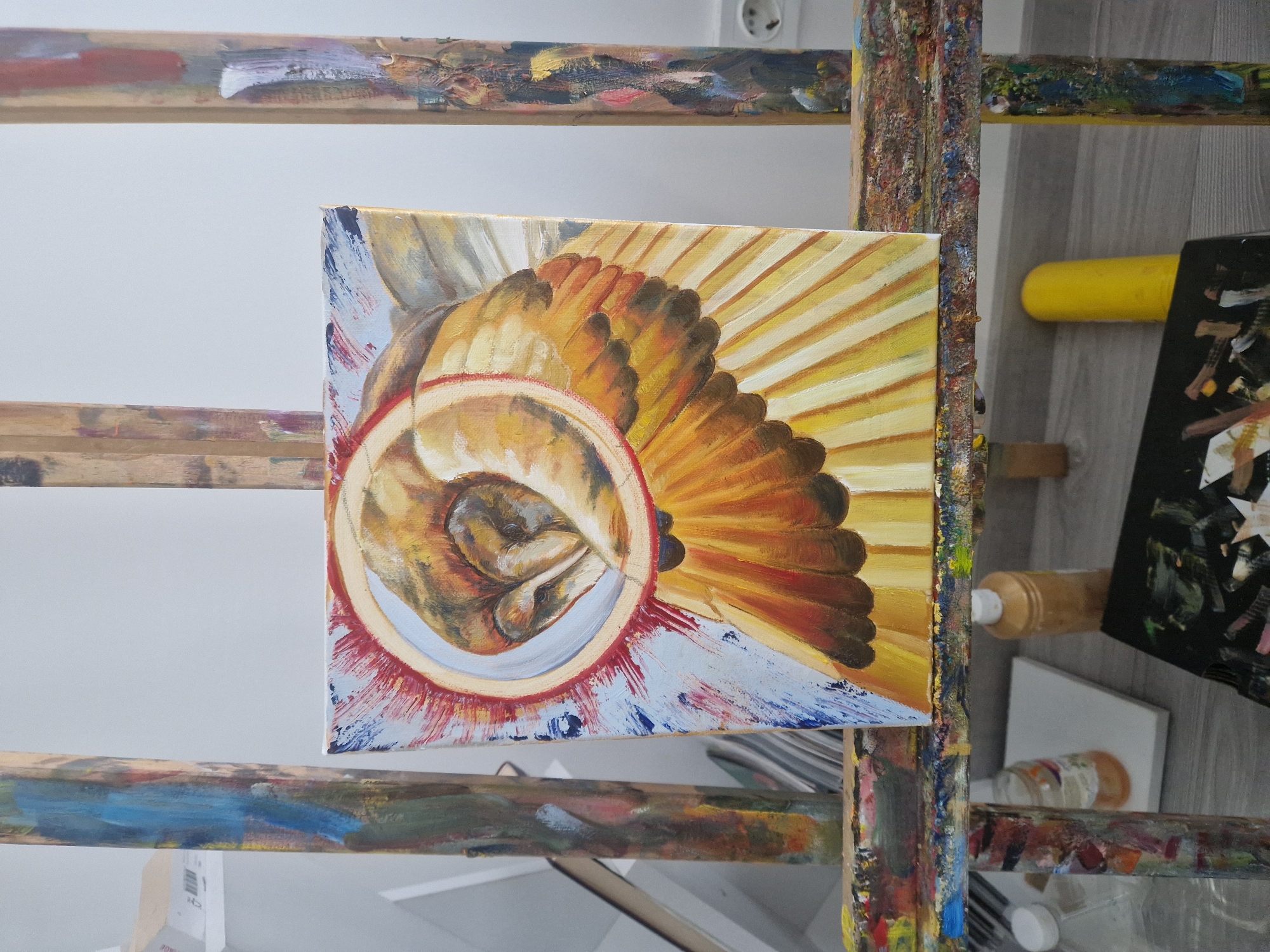 An art studio scene features a painting displayed on a wooden easel, which is heavily splattered with dried paint, indicating extensive use. The painting itself is a square canvas depicting a central, curled-up, human-like figure or embryo, rendered in warm brown and gold tones. This figure is encircled by a thick ring of white and red. Radiating outwards from this core are prominent, golden and reddish-brown, fan-like forms that resemble wings or feathers, creating a dynamic, almost spiritual aura. The background of the painting is a mix of light blue with expressive red and white brushstrokes.
The easel stands against a plain, light-colored wall, upon which a white electrical outlet is visible in the upper right. The floor appears to be made of light grey or wooden panels. In the background, to the right of the easel, various art supplies are visible, including a bright yellow cylindrical object, several bottles or jars of paint, and smaller canvases or boards. The lighting is bright and even, suggesting an indoor setting during the day or under artificial light. No legible text is visible on any of the objects within the frame.
