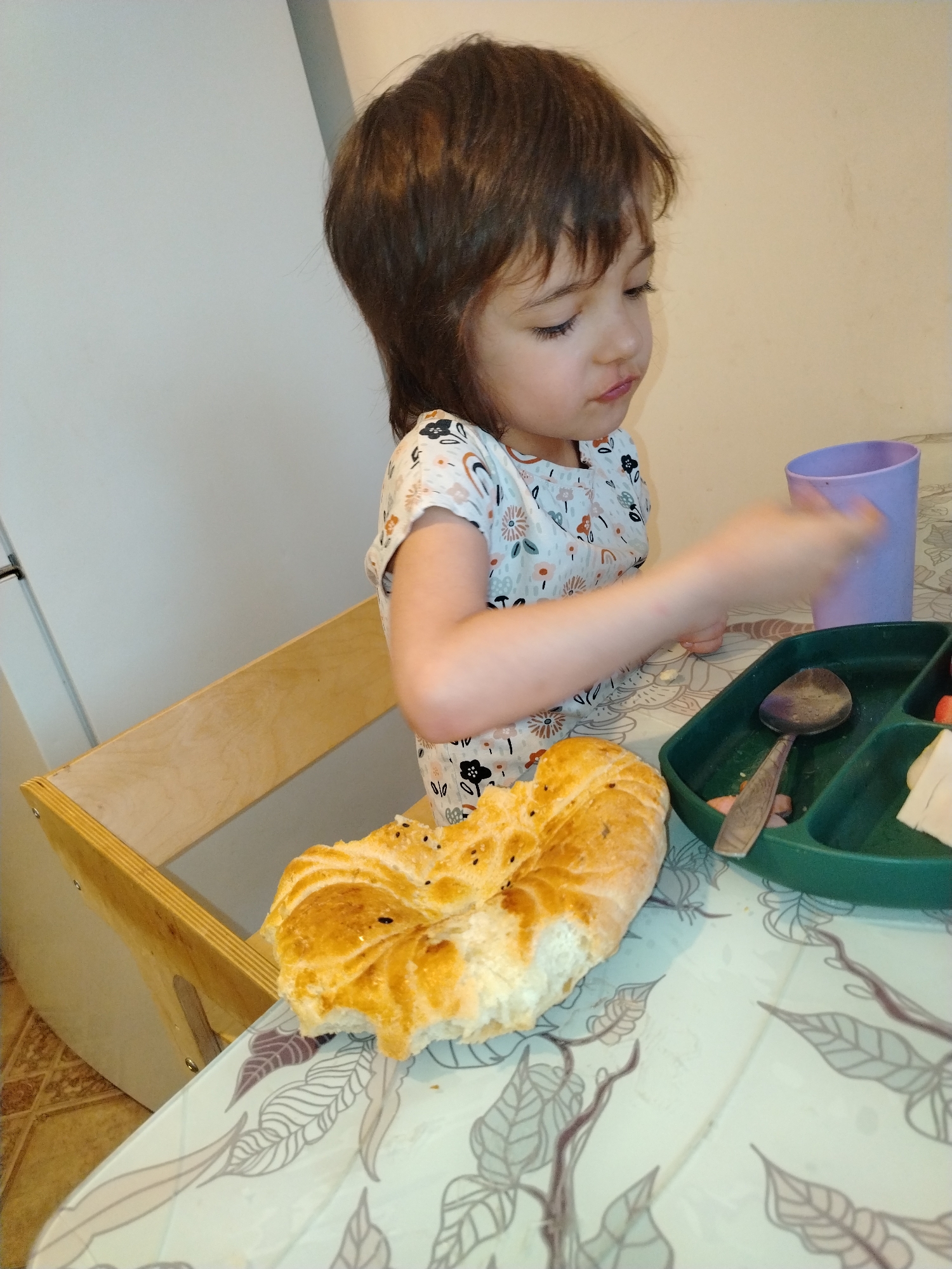 A young child sits at a table in an indoor setting. The child has short brown hair and is wearing a light-colored shirt with a floral pattern. In front of the child on the table, there is a piece of bread, a green divided plate with a spoon and some food items, and a purple cup. The table has a light-colored floral patterned surface. The child is reaching towards the cup. A wooden chair is visible to the left of the child. Based on available information, the location is potentially the city of Noviy, Russia.