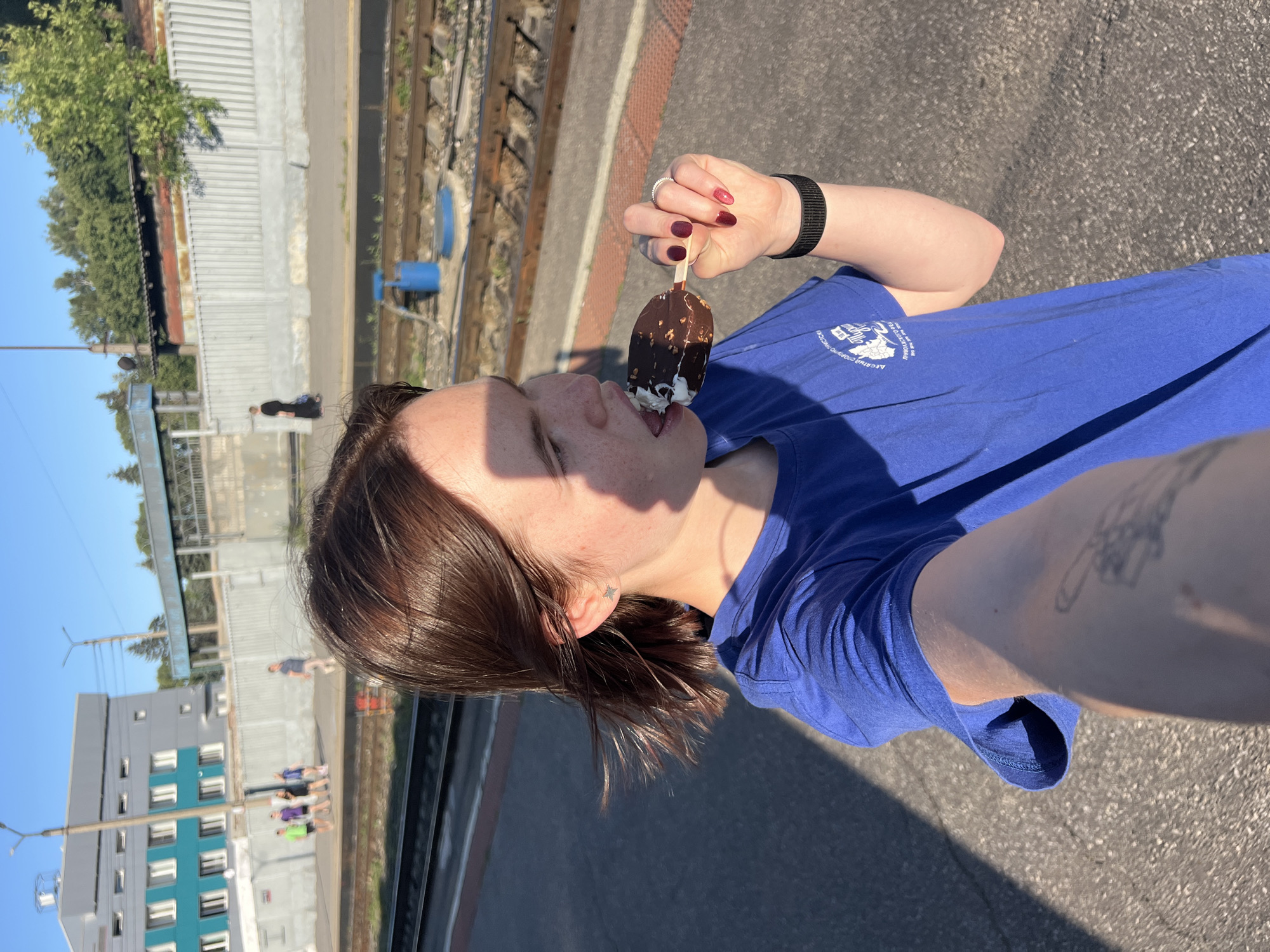 The media file depicts a young person enjoying an ice cream bar at a train station. The subject, with their hair in a messy bob, is shown from the chest up, wearing a blue t-shirt with a white graphic logo. They are holding the ice cream bar with their right hand, which has dark red nail polish. A black fitness tracker is visible on their left wrist. The person's face is partially obscured by a shadow from the sun, but their mouth is open as they lick the ice cream. A small star-shaped earring is visible in their left ear.
The background suggests an outdoor setting at a train station, with train tracks and a concrete platform visible. In the distance, there's a multi-story building with a teal and grey facade, and other people can be seen on the platform. The lighting indicates it's daytime, likely sunny. The overall mood appears casual and pleasant. The logo on the t-shirt is partially visible and appears to have text in Russian and a graphic element.