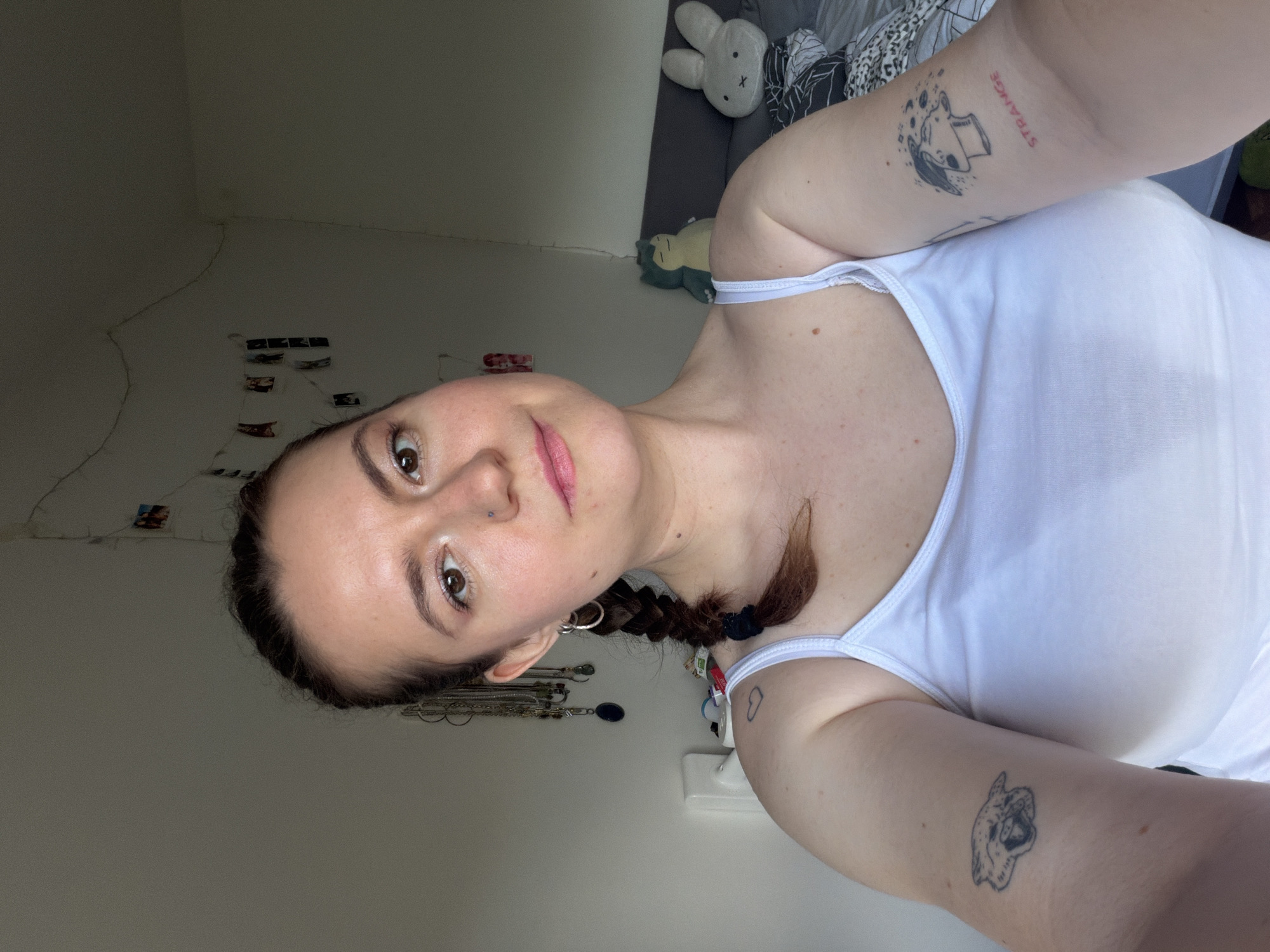A young woman with dark hair, styled in a single braid over her left shoulder, looks directly at the camera with a neutral expression and a slight smile. She has brown eyes, a labret piercing, and is wearing a white tank top.
She has several tattoos on her arms. On her right upper arm, a tattoo depicts a stylized head with a book on top, surrounded by abstract shapes. Next to this, the word "STRANGE" is tattooed vertically in a reddish hue. On her left upper arm, a tattoo of a dog's head, possibly a bulldog, is visible, along with a small black outline of a heart closer to her shoulder.
The setting appears to be an indoor room, likely a bedroom. In the background to her right (viewer's left), a white plush bunny and a green plush toy are visible on a bed with patterned bedding. To her left (viewer's right), several small printed photos are casually taped to a light-colored wall, accompanied by what looks like string lights or a wire. Below her left arm, a collection of necklaces and other jewelry hangs against the wall, and a white electrical outlet is also visible. The lighting is soft and ambient, suggesting an indoor environment without harsh direct light. The woman appears to be taking a selfie or posing for a close-up photograph.