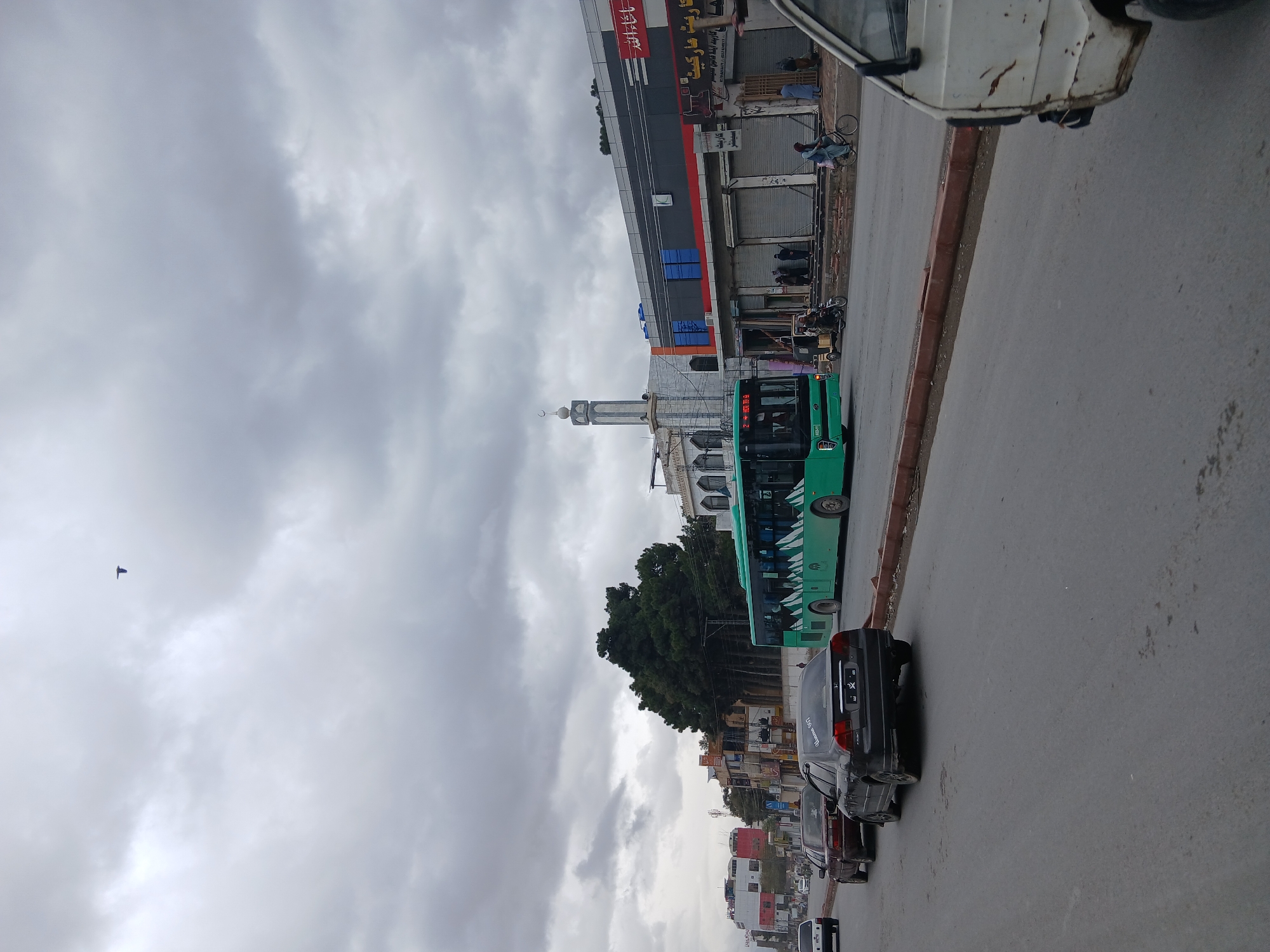 The image depicts a street scene in Quetta City Tehsil, Pakistan, under a cloudy sky. A green bus is visible on the right side of the road. Several cars are parked behind the bus. Buildings line the street. A mosque with a tall minaret is also seen in the background. People are present near the buildings along the street. A bird is flying in the sky.