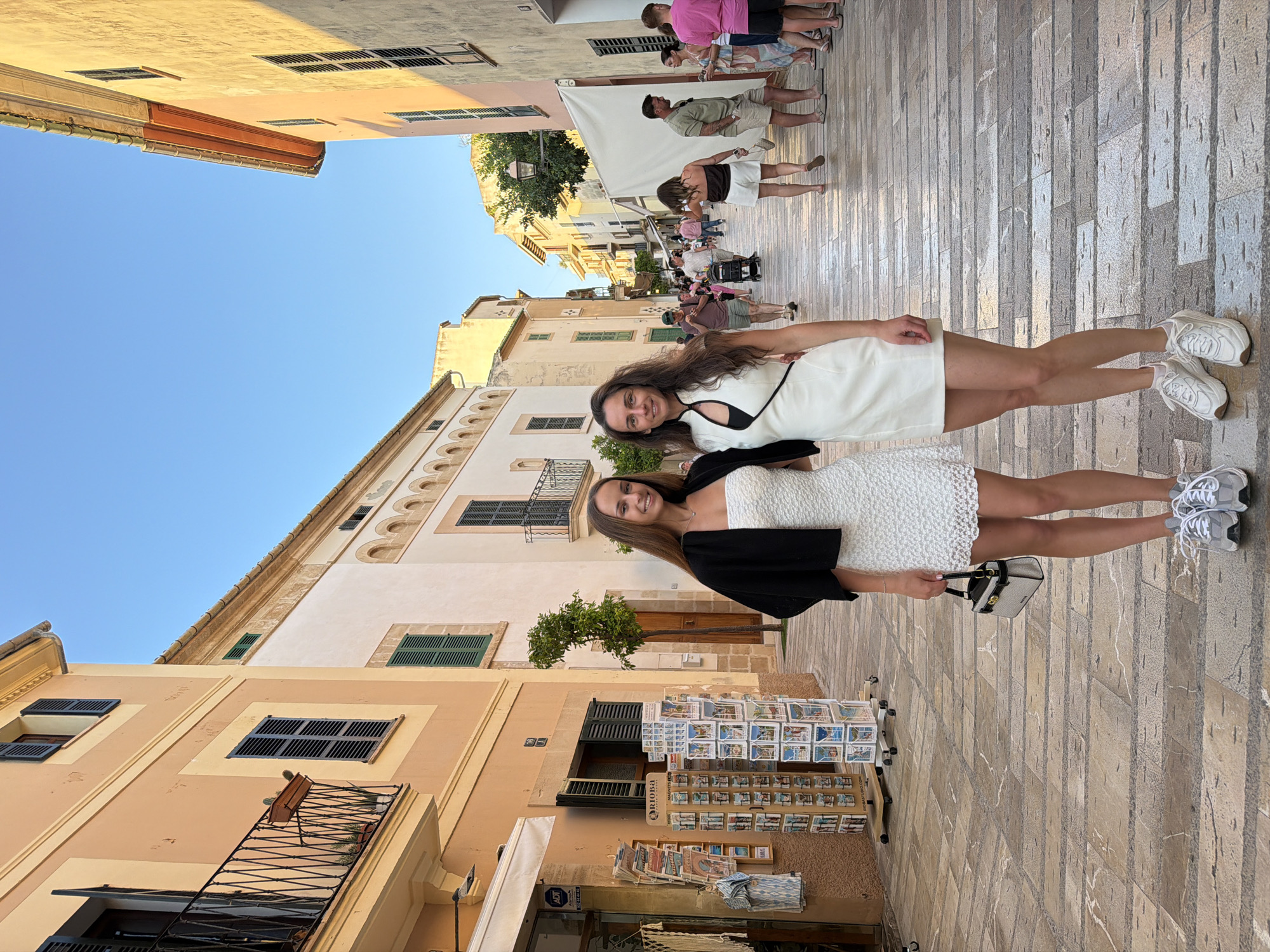 Two young women stand in a street in Alcúdia, Spain. The woman in the foreground wears a white dress, a black cardigan, and white sneakers. She carries a small purse. The other woman wears a white dress with black trim and white sneakers. Other people are walking in the background. The street is made of stone and is lined with buildings. A postcard stand is on the left. The sky is clear.