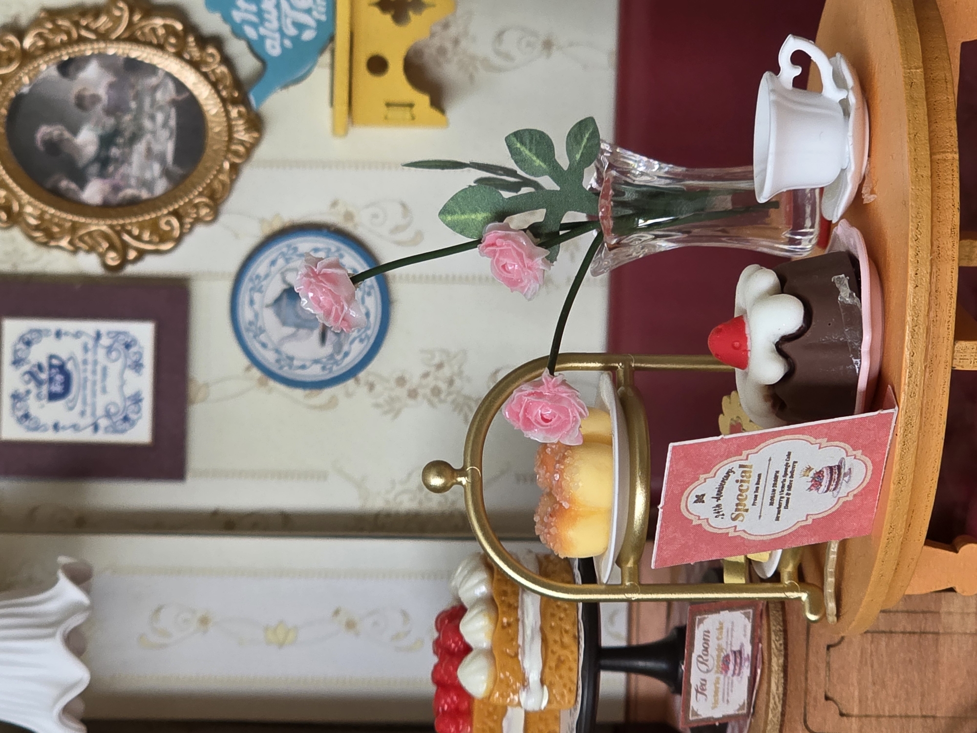 The image shows a miniature tea room setting. In the foreground, on an orange wooden table, are several miniature desserts. A chocolate cake with a white frosting and a red cherry on top sits on a pink plate. Next to it is a white teacup and saucer. To the left, a pink menu card is propped up, displaying "24th Anniversary Special" and details about strawberry Victoria sponge cake. Behind this, on a tiered golden stand, are miniature cakes. The top tier holds two small yellow cakes dusted with sugar, adorned with a pink rose. The bottom tier holds two larger layered cakes topped with red berries and white cream. To the far left, another menu card reads "Tea Room Victoria Sponge Cake." The background features dollhouse wallpaper with a floral pattern and framed pictures. A miniature blue teapot is visible on a yellow shelf. The scene is lit by a miniature lamp with a pleated white shade. The setting is a miniature representation of a tea room, not a real location in Baku, Azerbaijan.
