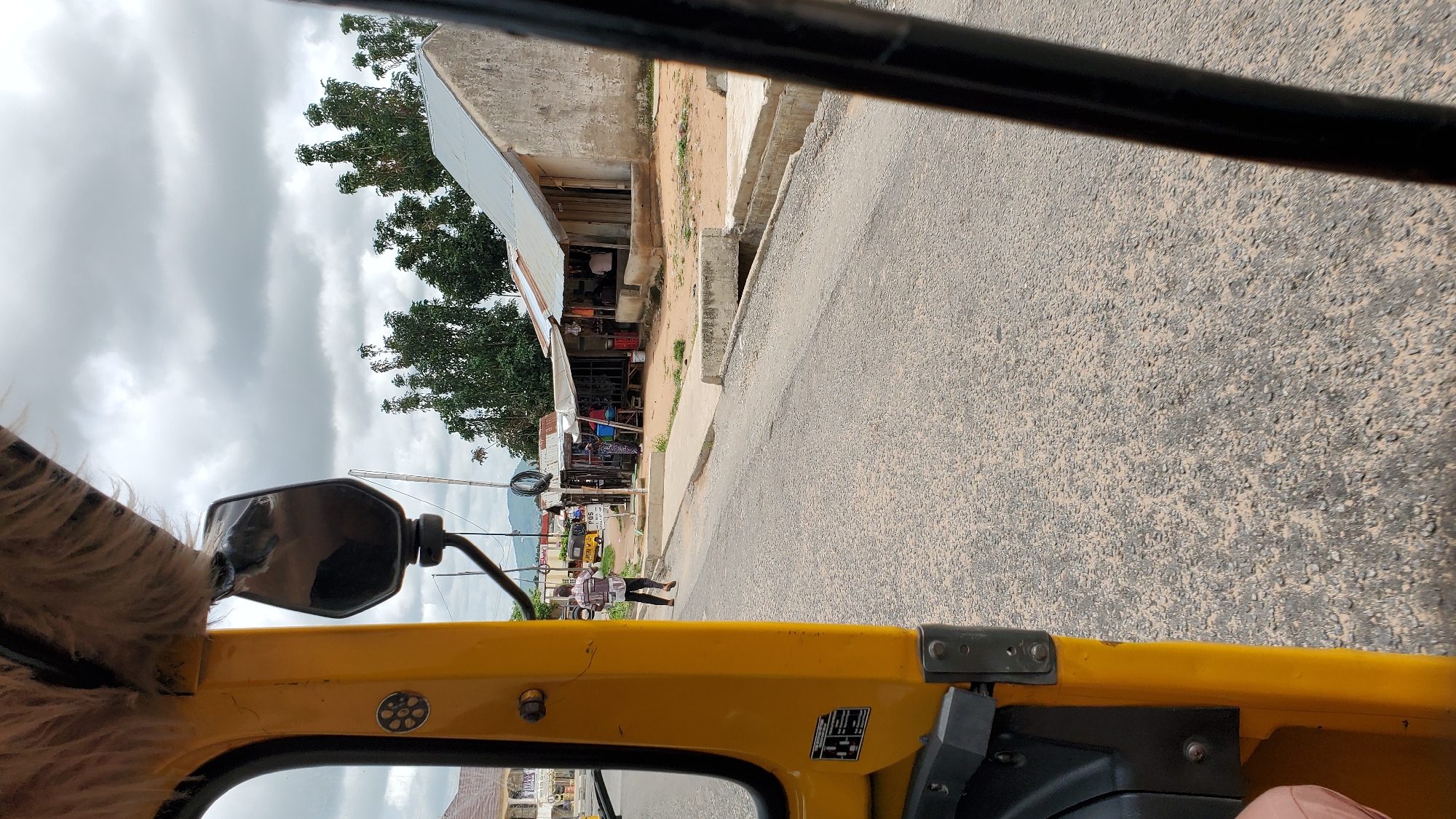 A person is walking on a dirt road next to a yellow vehicle. The person is wearing a white and brown patterned shirt and dark pants. In the background, there are several shops with corrugated metal roofs and a cloudy sky. The scene appears to be in Jalingo, Nigeria.