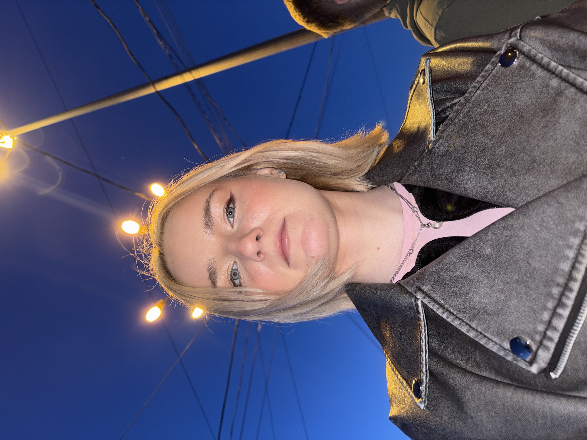 A young woman with blonde hair, wearing a pink shirt and a black leather jacket, poses for a selfie at night. She has a delicate silver necklace with a pendant, and her hair is styled with small lights clipped to it, resembling a halo. Her makeup includes winged eyeliner. The background is a deep blue night sky with visible power lines and a street lamp. The woman is in the foreground, looking directly at the camera. To her right, partially visible, is the shoulder and back of another person wearing a dark hoodie. The scene is likely in an outdoor urban setting.