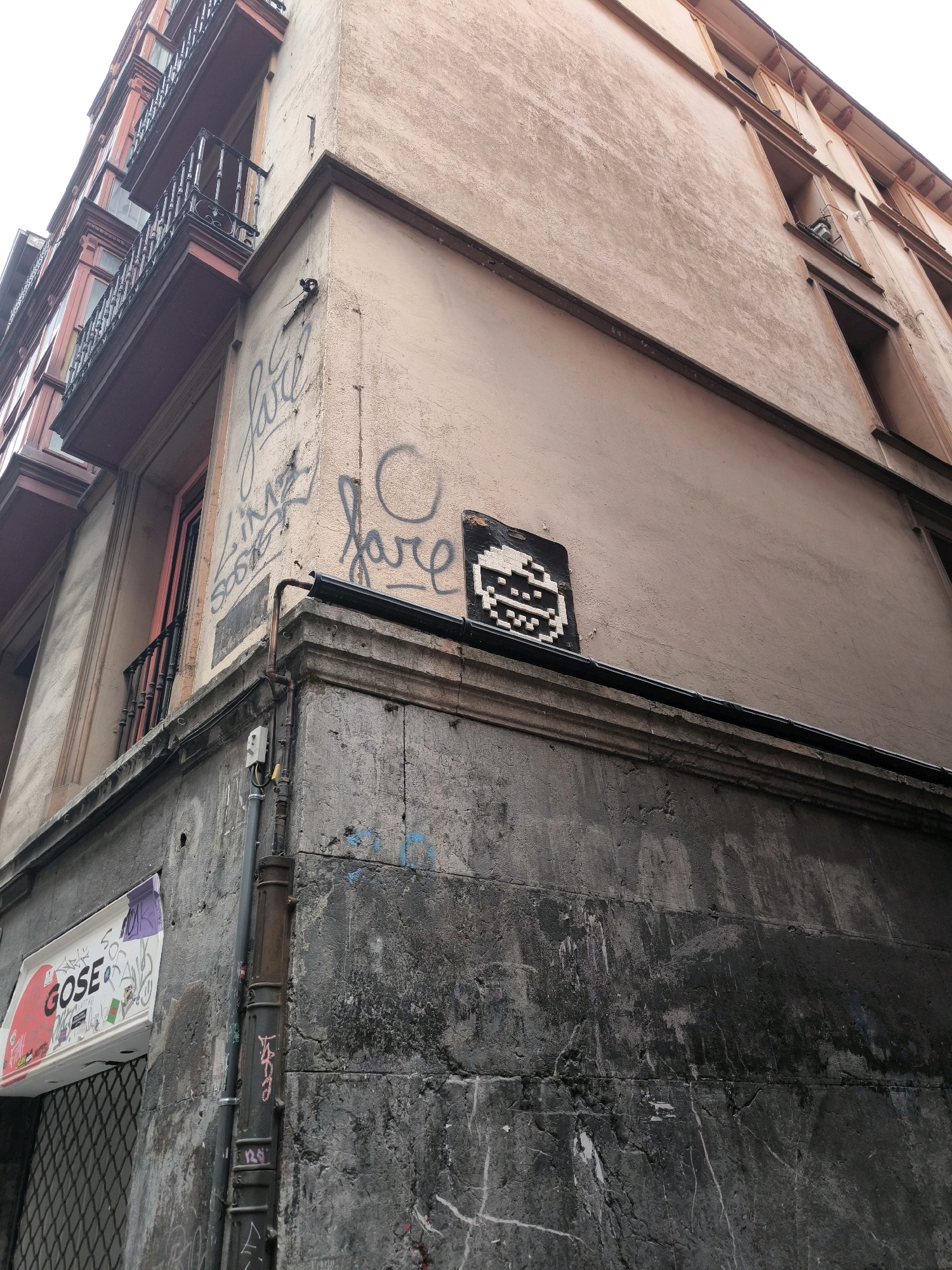 An outdoor, low-angle, medium shot captures the corner of a weathered, multi-story building in Bilbao, Spain. The building's facade is a faded peachy-pink on the upper floors and dark, mottled grey stone on the lower level, marked with various graffiti tags in black, grey, and red.
The upper portion of the building features balconies with dark metal railings and some red decorative elements. On the peachy-pink wall, a pixelated mosaic artwork of a smiley face, rendered in black and white, is affixed above a dark drainpipe. Below this, the lower section of the building presents a rough, textured stone surface. To the left, a sign with red and white lettering that reads "GOSE" is mounted, partially obscured by a chain-link fence. The overall scene is urban and shows signs of age and public art.