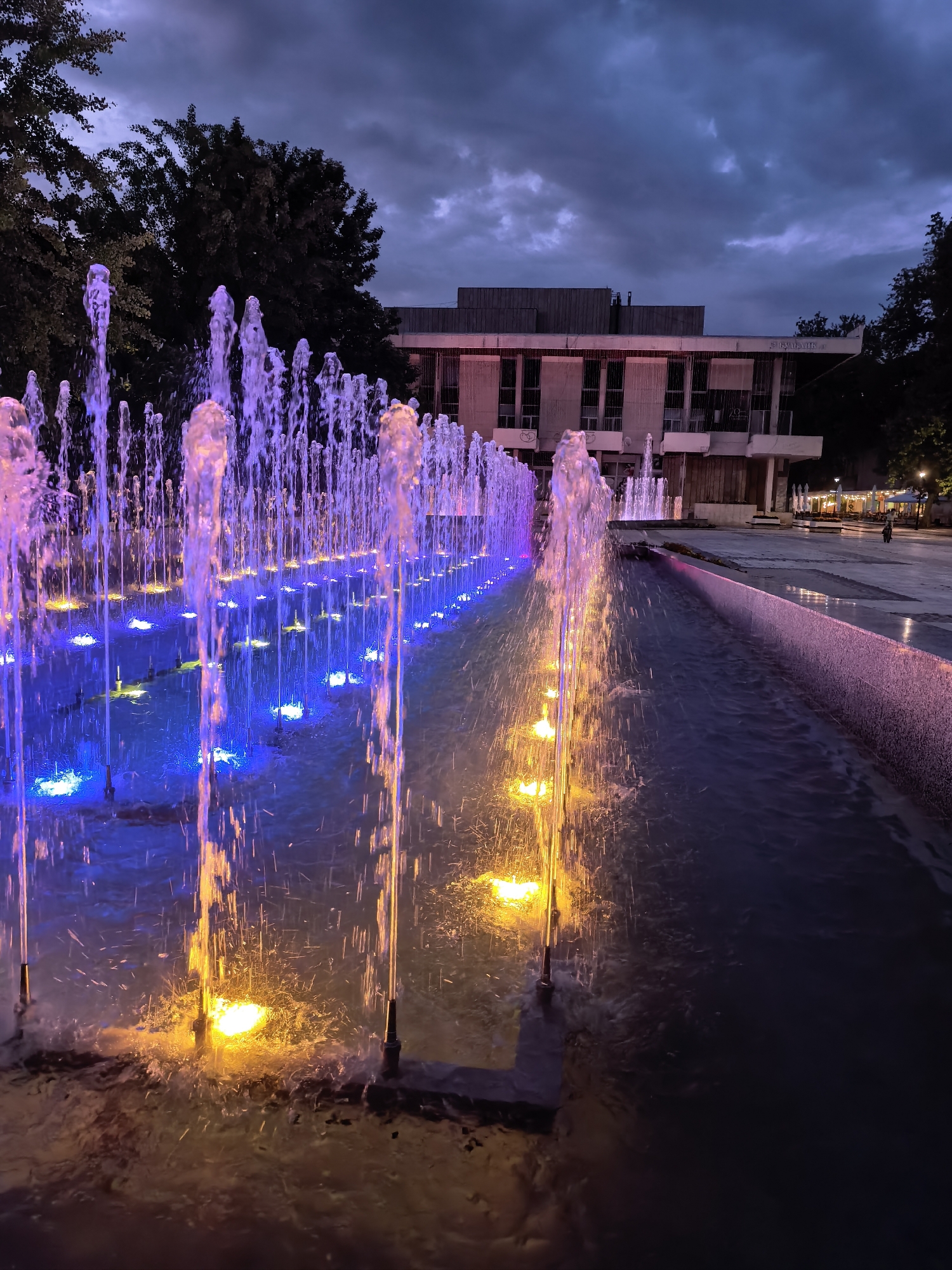 The image captures an outdoor urban scene at dusk or night, featuring a vibrant, illuminated water fountain system in the city of Svishtov, Bulgaria.
In the foreground and midground, a long, linear fountain runs from left to right, with numerous vertical water jets spraying upwards. The water jets are illuminated by colored lights embedded in the fountain basin. On the left side, the jets are predominantly purple/pink, transitioning through blue in the middle, and ending with yellow/orange on the right. The water flows into a narrow, dark channel alongside the main fountain.
In the background, positioned centrally, is a multi-story building with a flat roof, visible through and above the fountain jets. A sign on the building's facade, partially visible, reads "Народно читалище "Еленка и Кирил Д. Аврамови"" (National Community Centre "Elenka and Kiril D. Avramovi"), identifying the landmark in Svishtov. The building features multiple windows and a somewhat austere, likely concrete or stone, architectural style. To the left and right of the building, dark, dense trees are silhouetted against the twilight sky.
The sky above is overcast with dark, heavy clouds, indicating either a late evening hour or an approaching storm. Faint ambient light reflects off the clouds. To the far right, beyond the fountain and a paved plaza, an outdoor dining area with several white umbrellas and tables is visible, with indistinct human figures present in the distance. The ground immediately surrounding the fountain is a paved surface, part of a pedestrian area or public square. Artificial lighting from the fountain and distant streetlights is the primary light source in the scene.