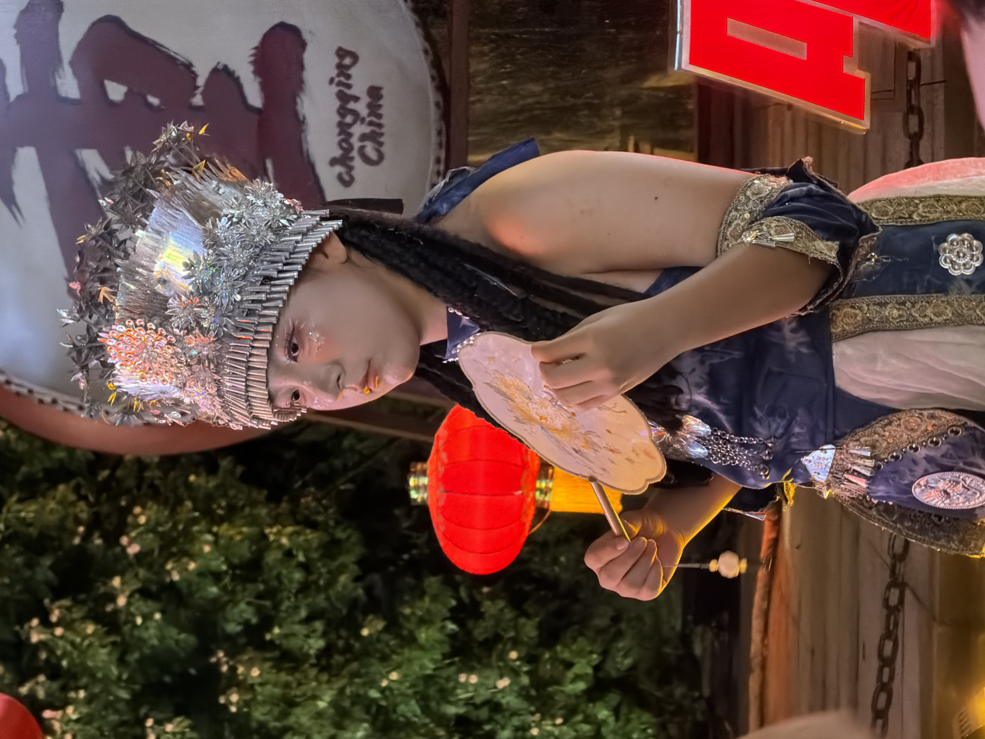 A young person wearing an ornate, silver, headdress adorned with glitter and reflective materials stands in front of a large, circular object with Chinese characters and "chongqing China" written on it. The person's face is painted with glitter on the cheeks and eyes. They are wearing a blue and white traditional costume with intricate gold embroidery and jewelry. In their right hand, they hold a decorated fan. To the left of the person, a red lantern with gold tassels is visible, and behind them, lush green foliage is present. To the right, a large red object with white lettering, possibly a sign, is partially visible. The scene appears to be outdoors at night, with artificial lighting illuminating the subjects and surroundings. The location is identified as Yuzhong District, China.