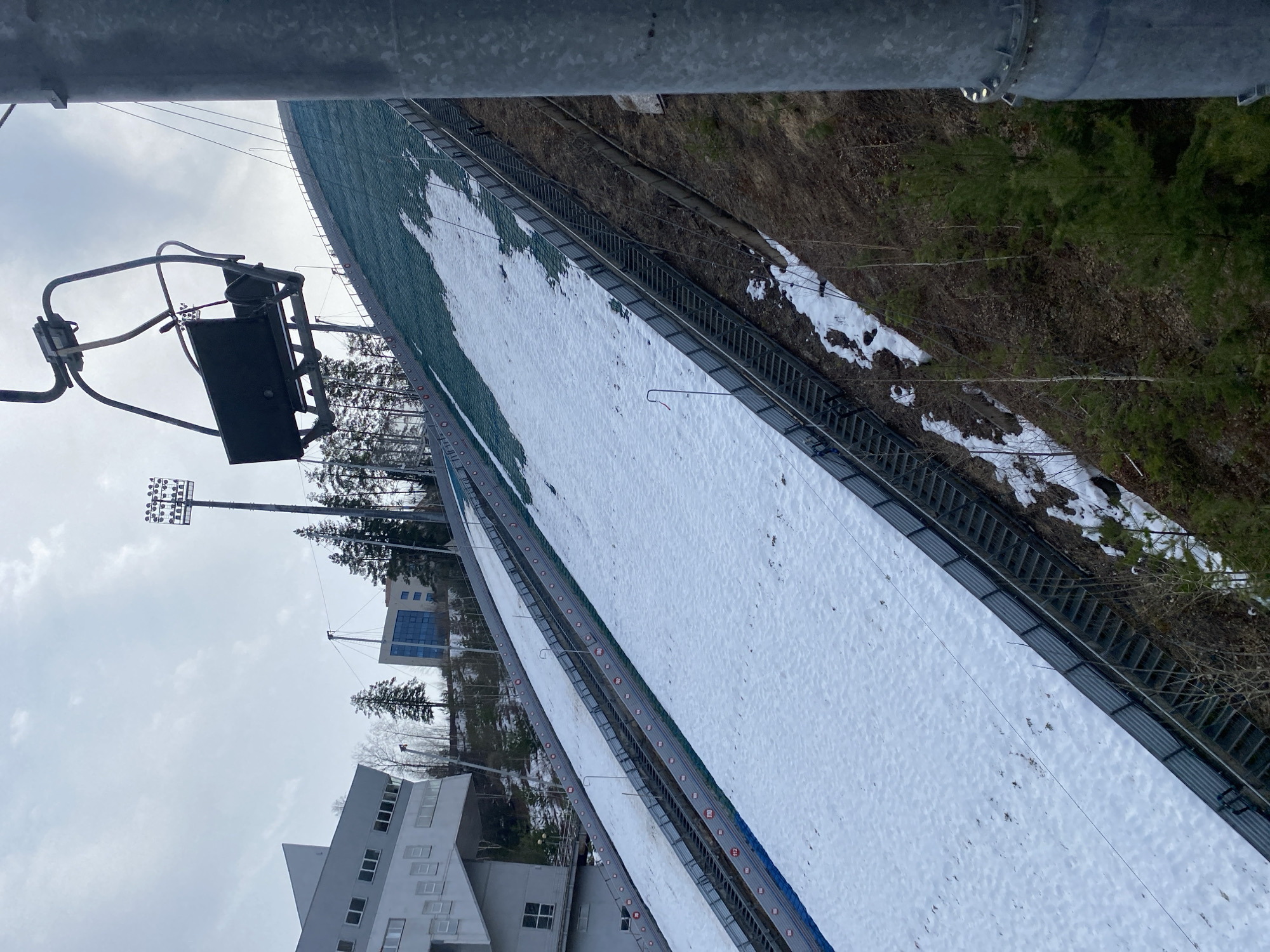 The image shows a snowy ski jump in Прикамский, Russia, viewed from a high vantage point, possibly a ski lift. A chairlift is visible in the foreground. The ski jump has a snow-covered track, safety fences, and distance markers. The surrounding environment includes trees, buildings, and a ski slope. Light towers are present. The sky is overcast.