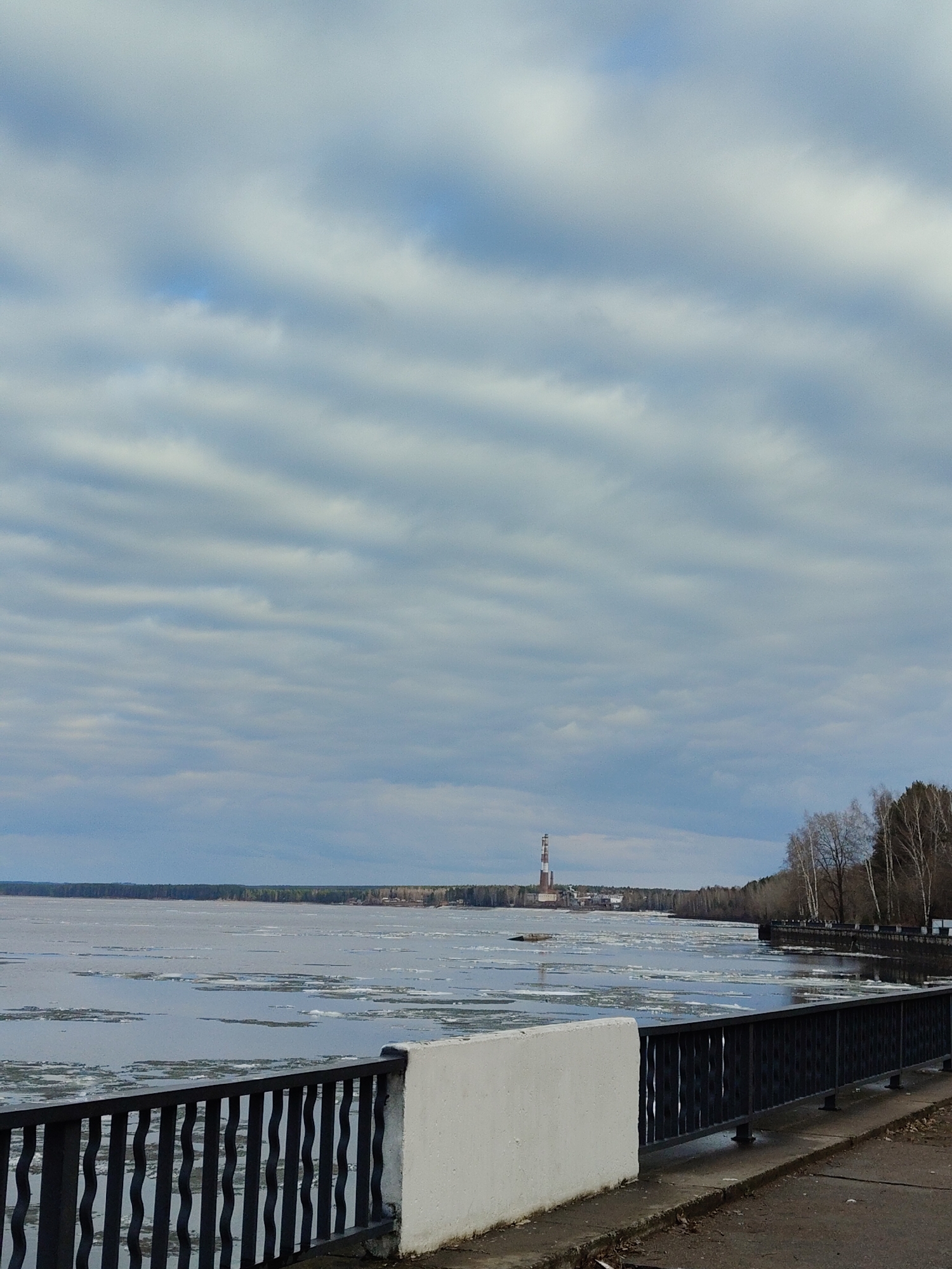 The image displays a wide, tranquil body of water, likely a river or large lake, partially covered with numerous melting ice floes. The scene suggests a transition from winter to spring, characterized by the ice breakup.
In the foreground, a robust black metal railing with wavy uprights extends across the lower right, bordering a concrete embankment. A white concrete wall or parapet is prominent in the immediate foreground, with a narrow concrete path visible below it.
The middle ground is dominated by the expansive water surface, dotted with floating chunks of ice. On the far bank, a distant treeline composed of both bare deciduous trees and some evergreen foliage stretches across the horizon. A prominent feature on the distant shore is a tall, slender industrial chimney or tower, distinctly marked with alternating red and white stripes. Several smaller industrial buildings are visible near its base. Closer to the right foreground, a group of bare-branched deciduous trees, possibly birch, lines the bank.
The sky is entirely overcast with layers of greyish-white clouds, giving the scene a soft, diffused light, indicating daytime. The weather appears cool or cold, consistent with the melting ice. There are no people visible, nor any discernible text. The overall atmosphere is quiet and observant of a natural process alongside an industrial presence.