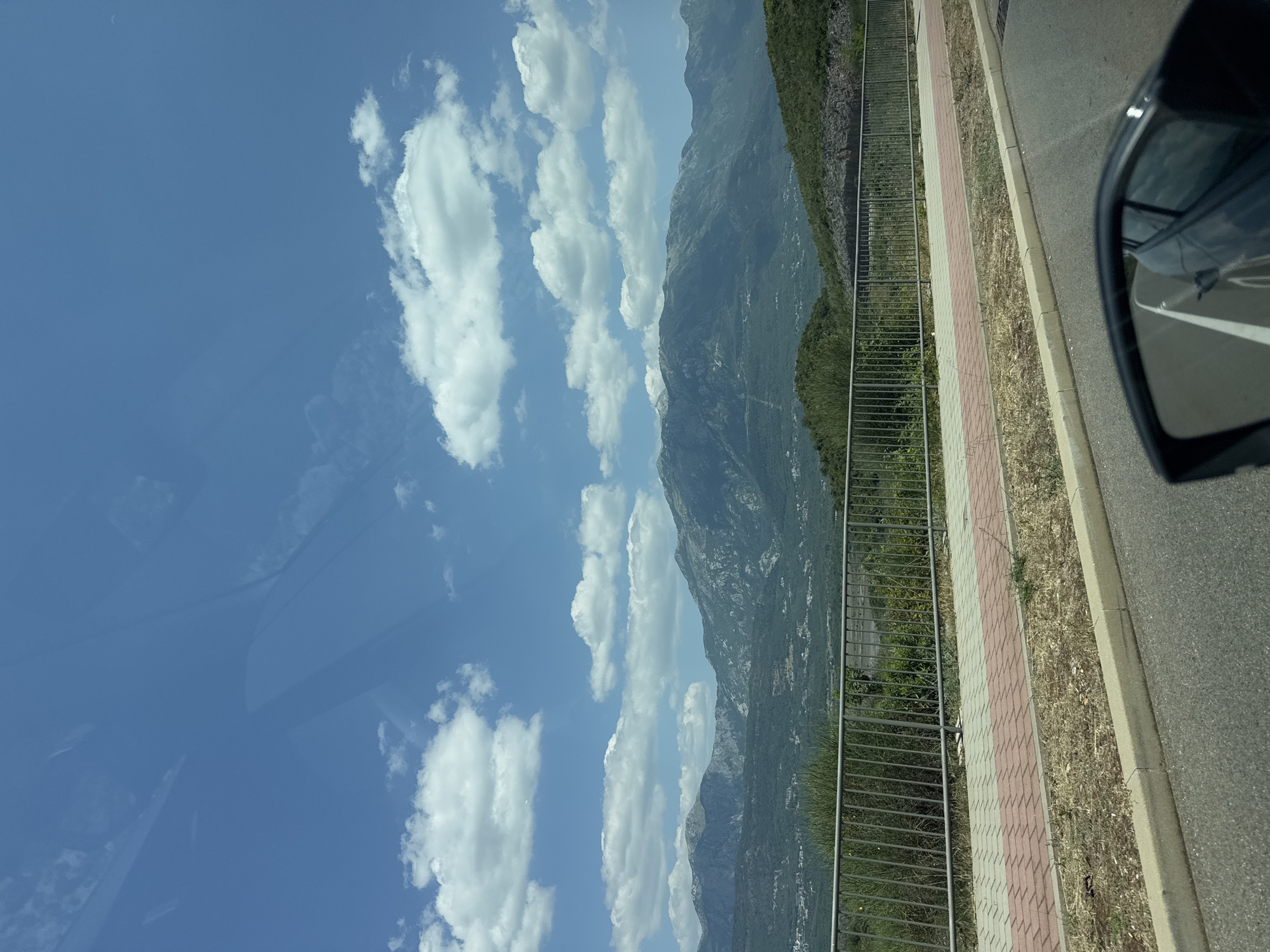 A bright, overcast day illuminates a scenic view of mountains and a coastline. The sky is a vibrant blue, dotted with fluffy white clouds. In the foreground, a paved walkway with red and grey brick patterns runs alongside a metal fence. Beyond the fence, a lush green landscape slopes down towards a distant, shimmering body of water, suggesting a coastal location. The rugged, verdant mountains dominate the background, their peaks partially obscured by the hazy atmosphere. The scene evokes a sense of natural beauty and tranquility.