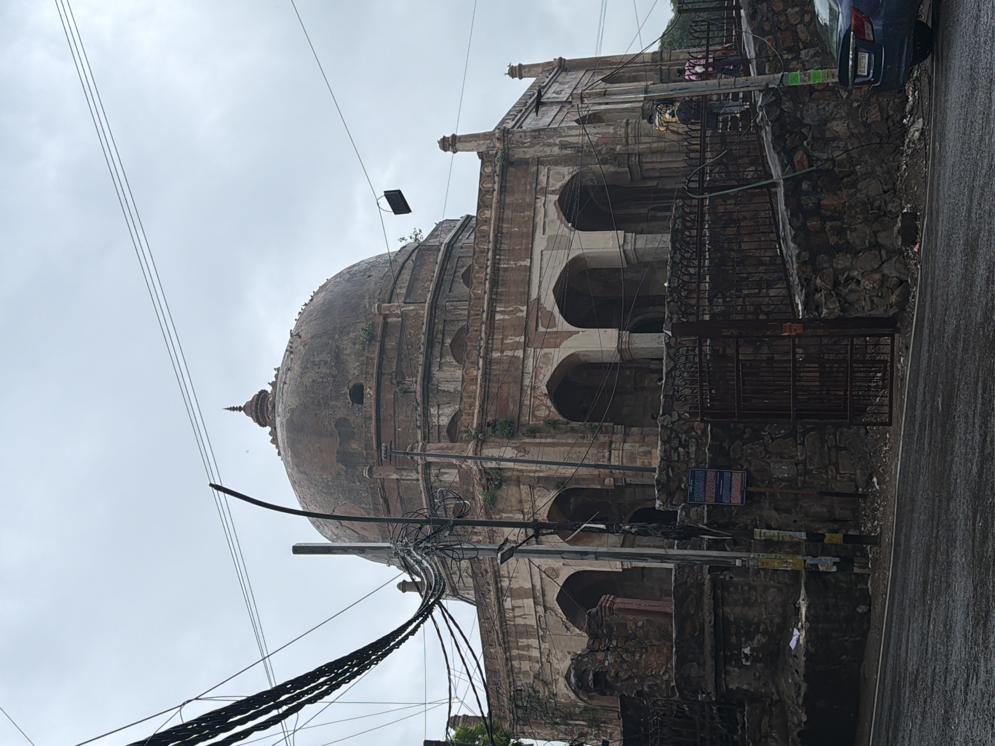 The image depicts a view of the tomb of Safdarjung in Delhi, India. The domed mausoleum, constructed of red sandstone and marble, is visible in the background, surrounded by overgrown vegetation. In the foreground, a stone wall with an iron gate separates the mausoleum from a paved road, which appears wet from rain. Electrical wires are prominently strung across the frame, obscuring parts of the sky. Several people are seen near the entrance to the tomb, and a blue car is parked on the side of the road. The overall atmosphere is overcast and slightly somber due to the weather.