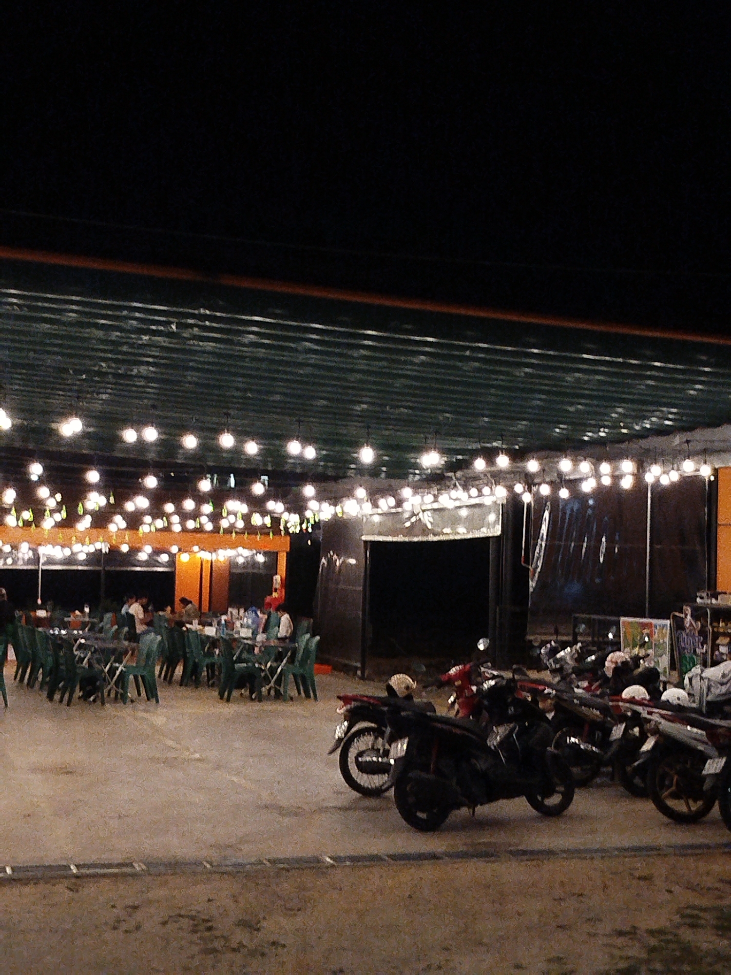 A night shot captures a restaurant scene in Da Lat, Vietnam, featuring an open-air dining area with tables and green chairs occupied by patrons. Overhead, numerous string lights illuminate the space, suspended beneath a corrugated metal roof structure.
To the right, several parked motorcycles add to the casual atmosphere. The flooring is a dark-toned concrete or similar material. The background suggests the presence of storefronts and advertising.