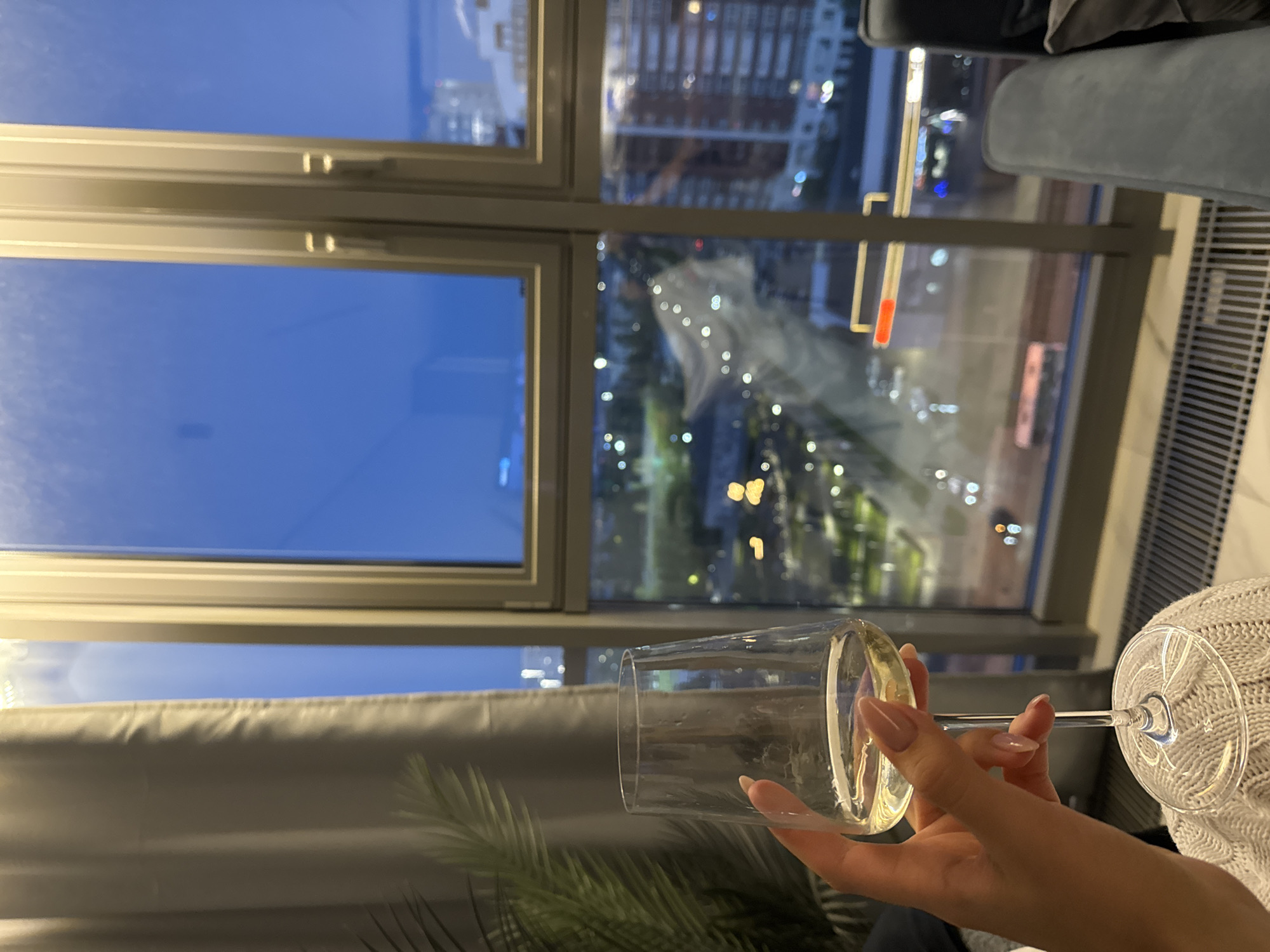 A slightly low-angle indoor shot depicts a hand holding a wine glass filled with a light-colored liquid. The hand has long, manicured fingernails with a pale pink color. The glass is partially obscuring a view of an urban setting through a window. The window frame is light-colored, and the view reveals a mix of natural and artificial light sources. There are buildings visible in the background, along with street lights and hints of greenery. The sky visible through the window is dark blue, indicating either evening or night. The bottom of the frame includes a glimpse of a green plant and a white knitted garment or blanket. There's no information about the location.