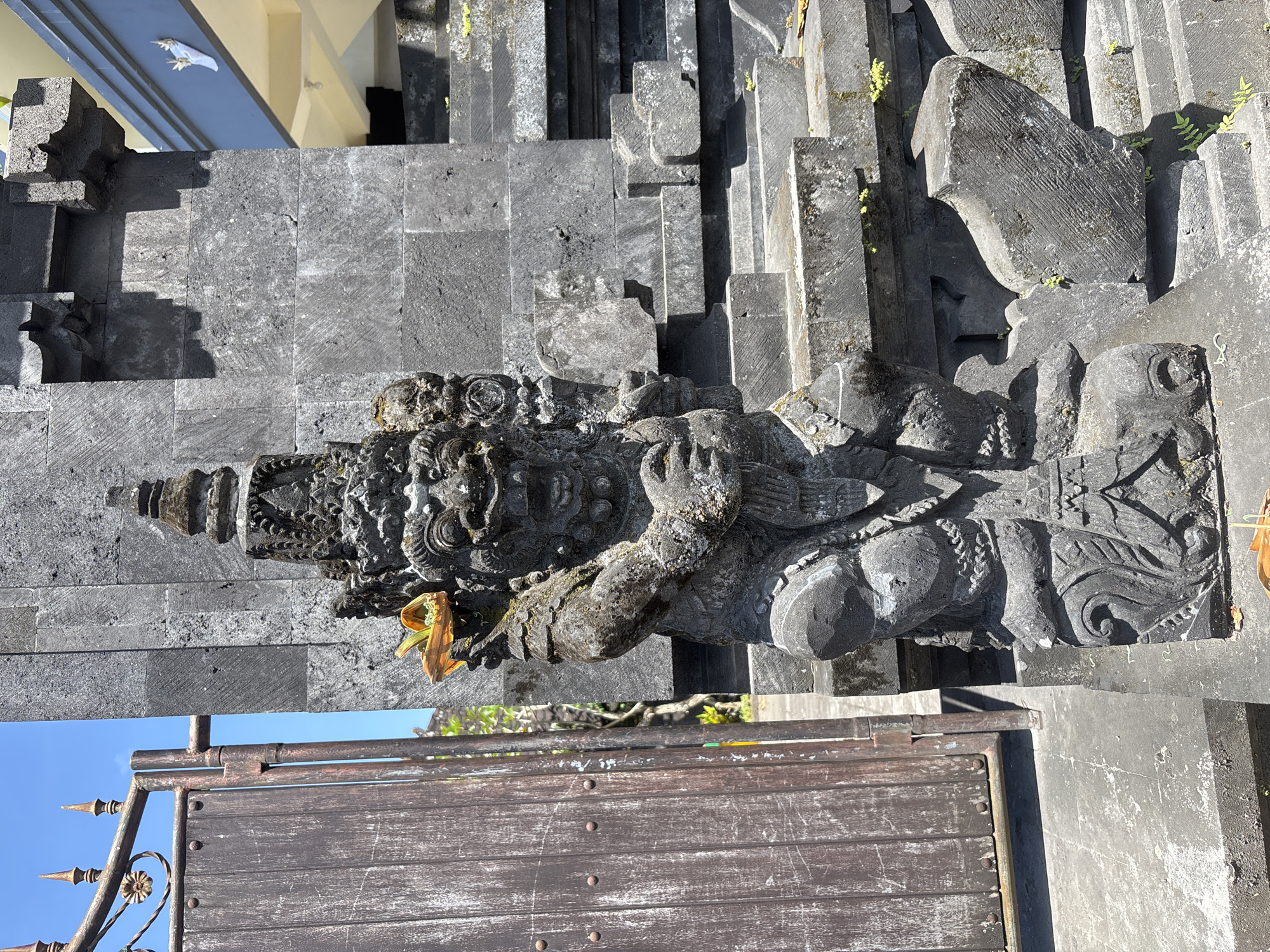 The image depicts a stone sculpture of a deity or mythical figure located at a temple in Kuta Selatan, Indonesia. The statue, carved from dark gray stone, is intricately detailed with facial features, ornate headdress, and stylized clothing. A yellow flower or offering adorns the sculpture. The sculpture is positioned against a background of gray stone steps and architectural elements, hinting at a temple complex. A section of a weathered wooden gate with metal detailing is visible in the foreground. The weather appears sunny, casting shadows on the stone surfaces.
