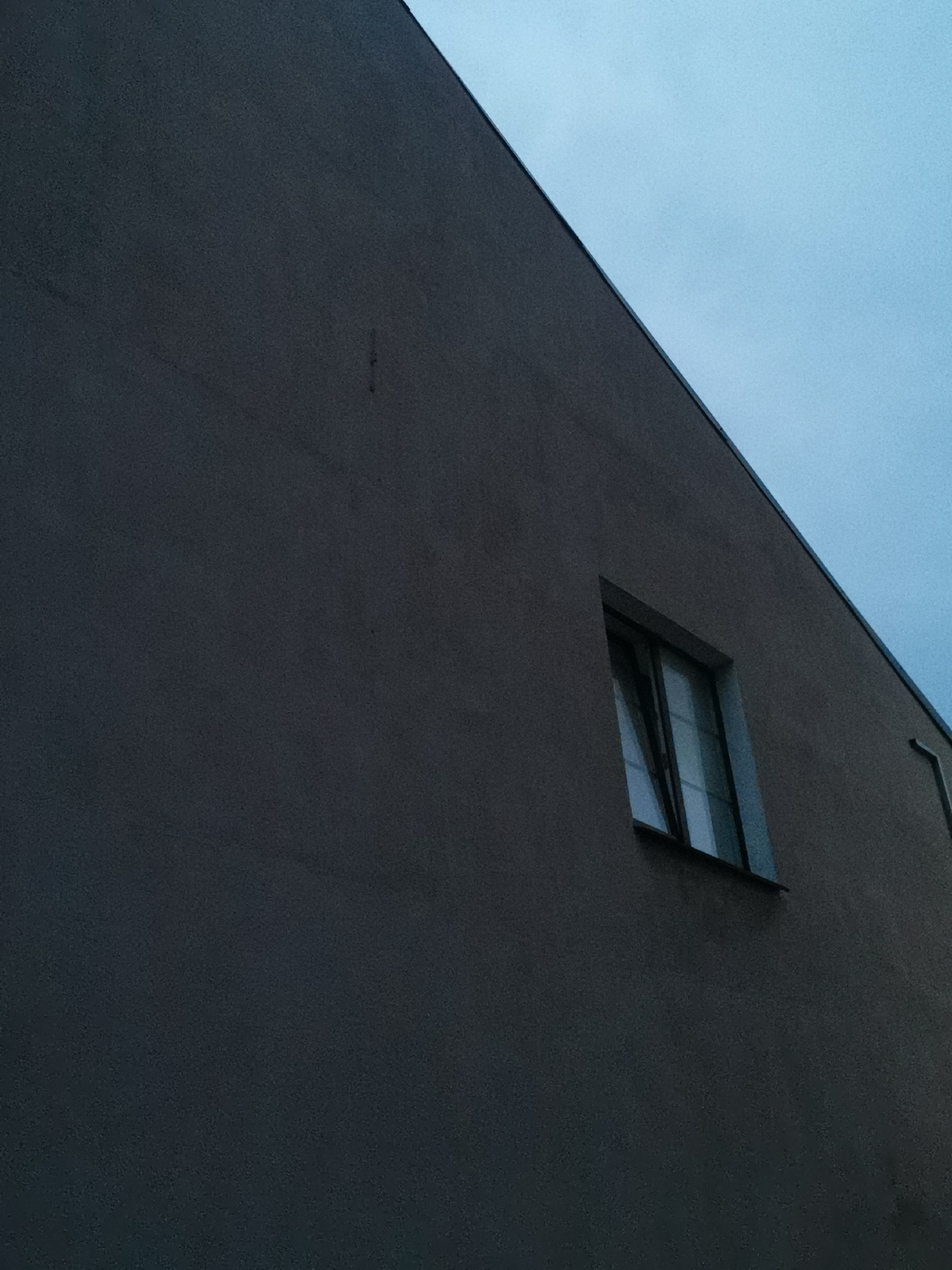 The image shows the exterior of a dark gray building under a slightly overcast, light blue sky. A single, small rectangular window is visible on the building's facade, slightly ajar. The window is divided into panes by thin, dark mullions and reflects the sky. The building's wall has a textured surface. To the right of the window, a thin, vertical downspout is partially visible. The perspective is from below, looking up at an angle towards the sky. There are no people or any signs of life depicted in the image. The setting appears to be urban, possibly a residential or commercial building.