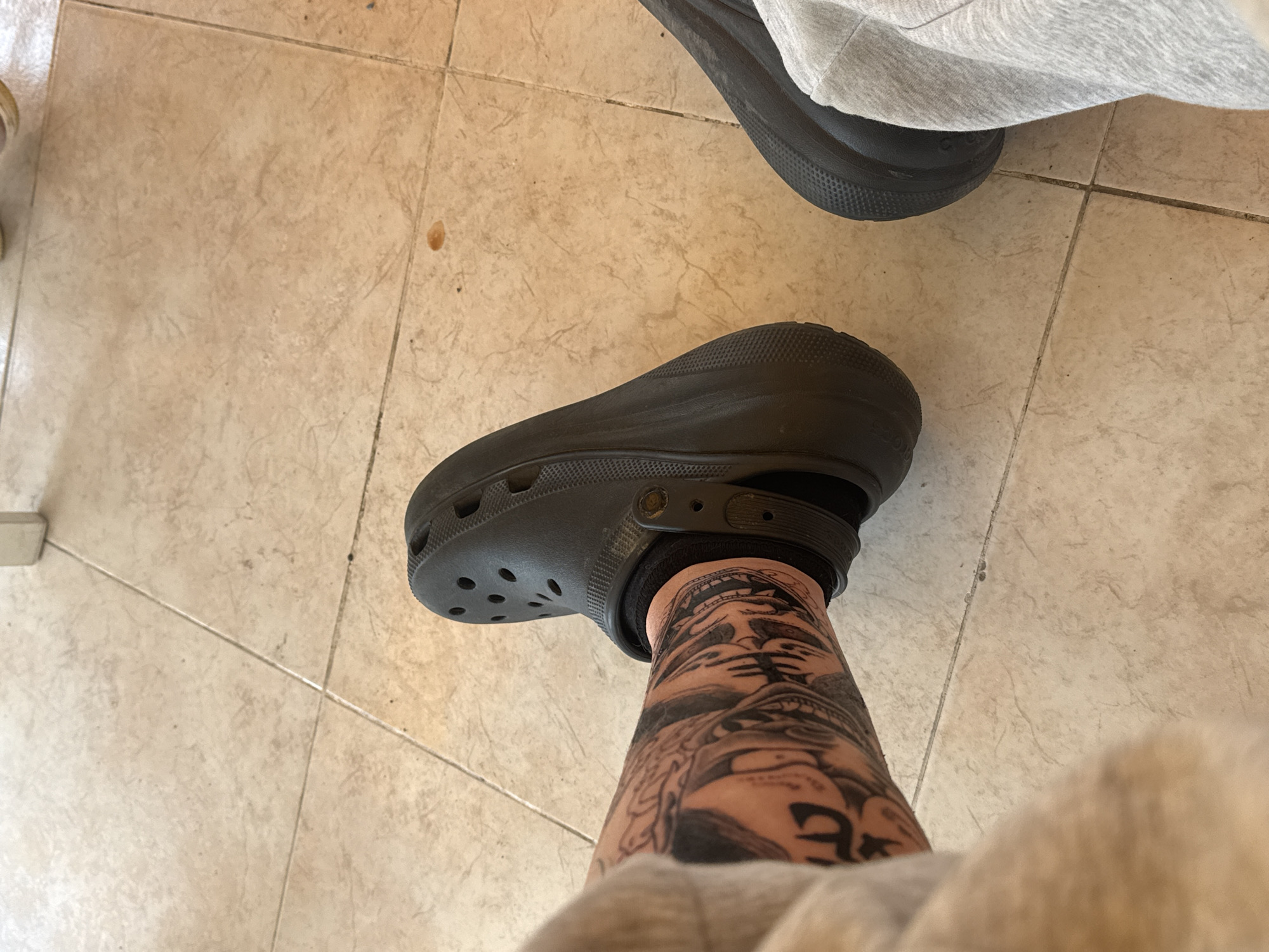A first-person perspective image shows a person's leg wearing a black Croc-style shoe and a black sock. The leg has a prominent black and gray tattoo extending upwards from the ankle. The person is wearing light gray sweatpants. Another black shoe, partially visible, is in the upper right of the frame, partially obscured by gray fabric. The background is a tiled floor with light brown tiles. There is no information to suggest that this photo was taken in Burgas, Bulgaria.