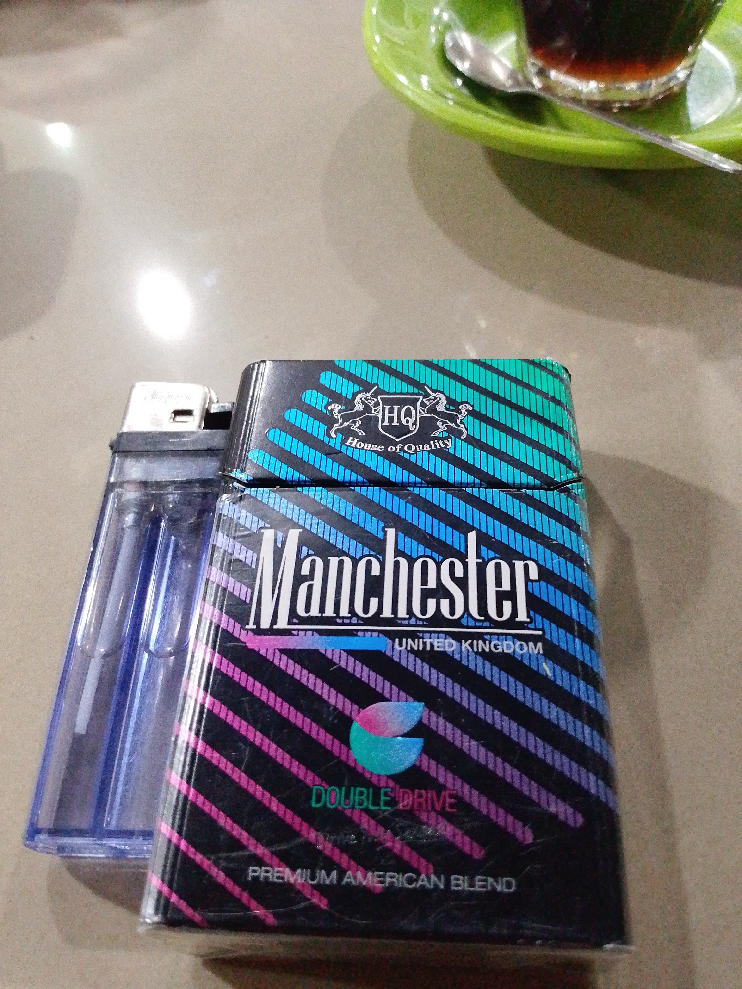 A pack of Manchester Double Drive cigarettes rests on a beige surface alongside a blue lighter. Above the cigarette pack is a green plate with a cup of dark liquid and a spoon. The pack's design features diagonal stripes of varying colors, including blue, pink, and black, with the brand name "Manchester" and "United Kingdom" printed on it. The cigarettes are identified as a "Premium American Blend". The image appears to be taken on a table, possibly in a café or restaurant. There is no indication of Lhokseumawe, Indonesia, beyond potentially the general stylistic choices of the setting.