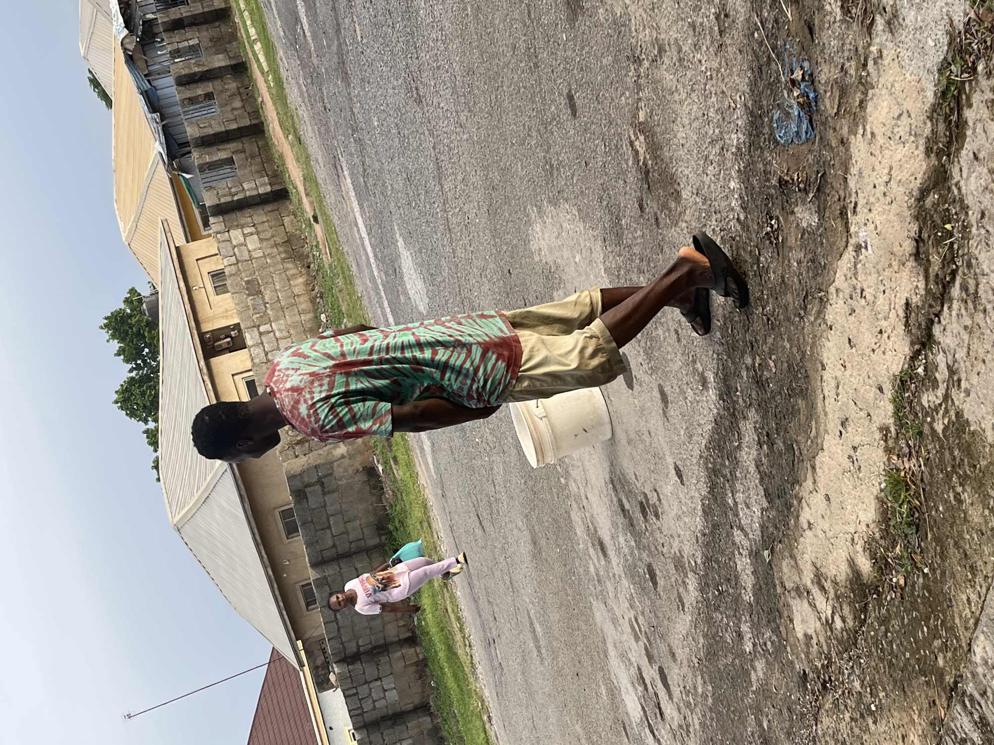 In Abuja, Nigeria, on a paved road with some scattered debris and puddles, a young man wearing a tie-dye t-shirt and khaki shorts walks away from the camera, looking over his left shoulder. Beside him, a white bucket sits on the ground. Further down the road, a woman in a pink t-shirt and pink pants carries a light blue bag and walks towards the camera. Behind them, a row of low-rise buildings with concrete block walls and corrugated metal roofs line the street. The sky is overcast.