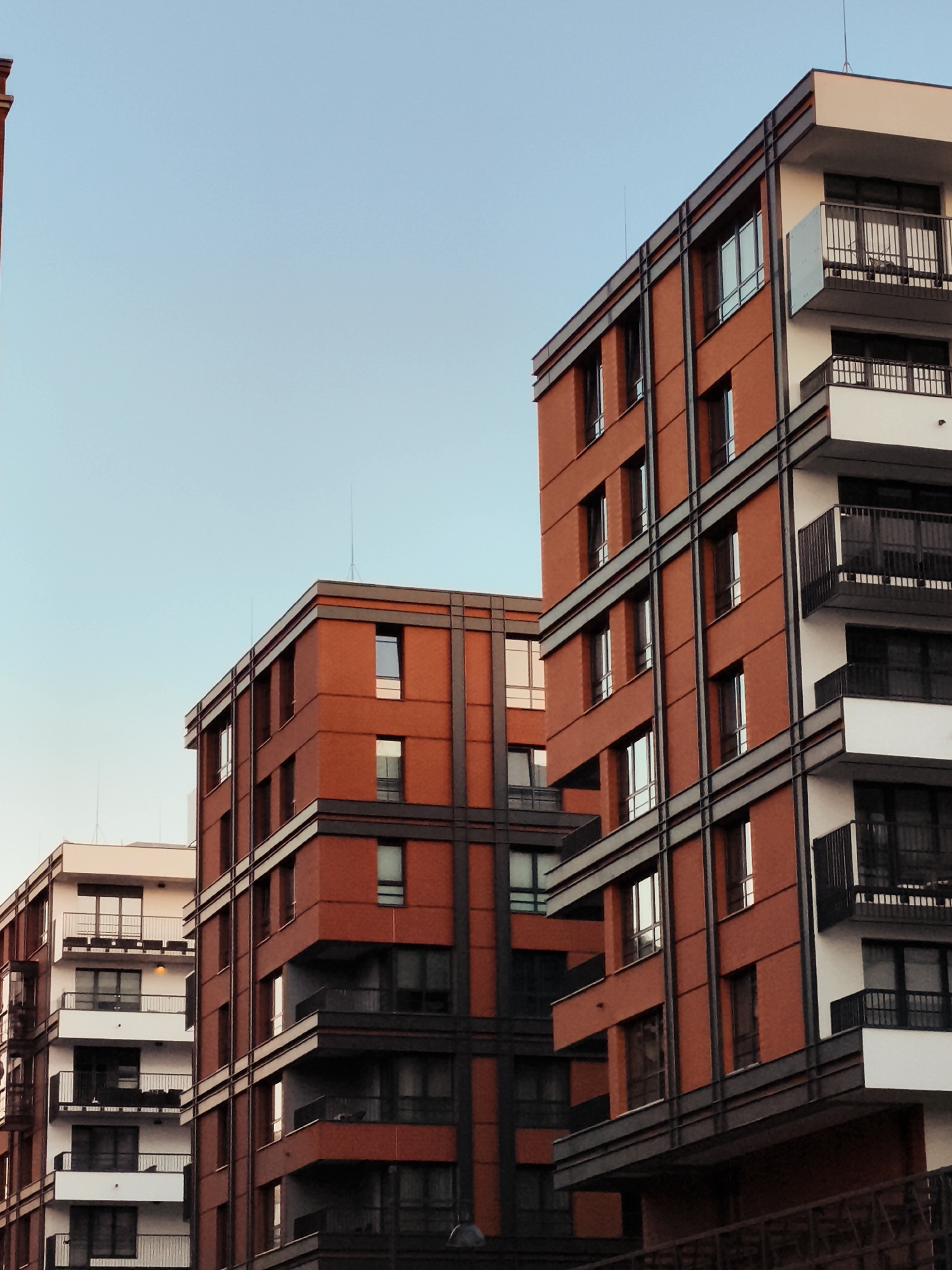 The image shows a close-up, angled view of several modern apartment buildings in Gdańsk, Poland, against a clear, pale blue sky. The buildings are primarily constructed with red brick and white facades, accented with dark grey horizontal and vertical framing elements. Multiple balconies with black railings are visible on the white sections of the buildings. The architecture is characterized by sharp lines and a geometric design. The lighting suggests it is either early morning or late afternoon, casting subtle shadows and highlighting the building materials. No people or specific activities are discernible in the scene.