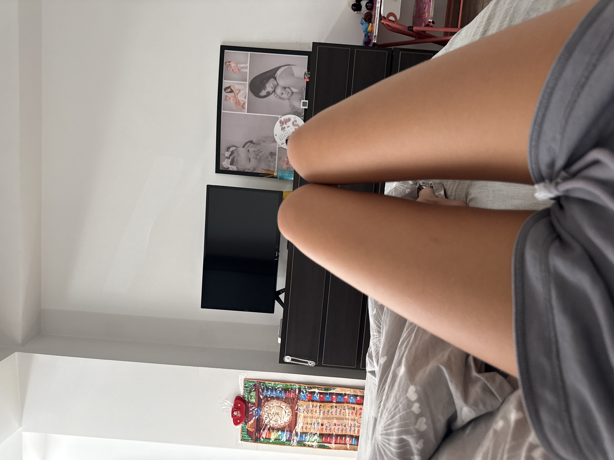 The image shows a person lying on a bed, facing away from the camera. The person's legs and upper thighs are visible, covered by grey shorts. The background shows a bedroom interior with a dresser, a television, a framed collage of photographs, and a decorative wall calendar. A small pink metal shelf unit is visible on the right side of the image. The scene is lit by natural light, suggesting it is daytime. The location is not specified in the image or accompanying text.