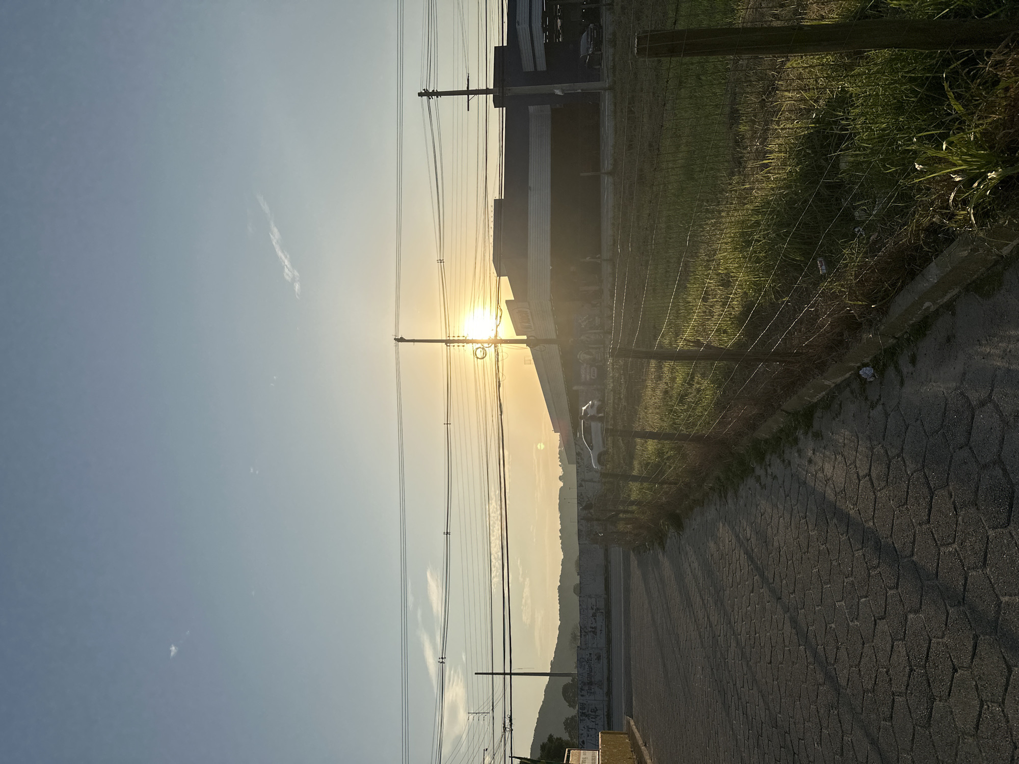 An outdoor, ground-level, medium shot depicts a paved walkway adjacent to a grassy field enclosed by a wire fence. The sun is setting, casting long shadows from the fence poles and overhead electrical wires across the walkway. In the background, a low-rise building with a dark facade is visible, possibly a commercial establishment, with a white car parked in front. Beyond the building, a line of hills is silhouetted against the bright sky. The overall impression is one of a somewhat urbanized natural landscape during dusk. This scene is likely located in Florianópolis, Brazil, given the context of the prompt.