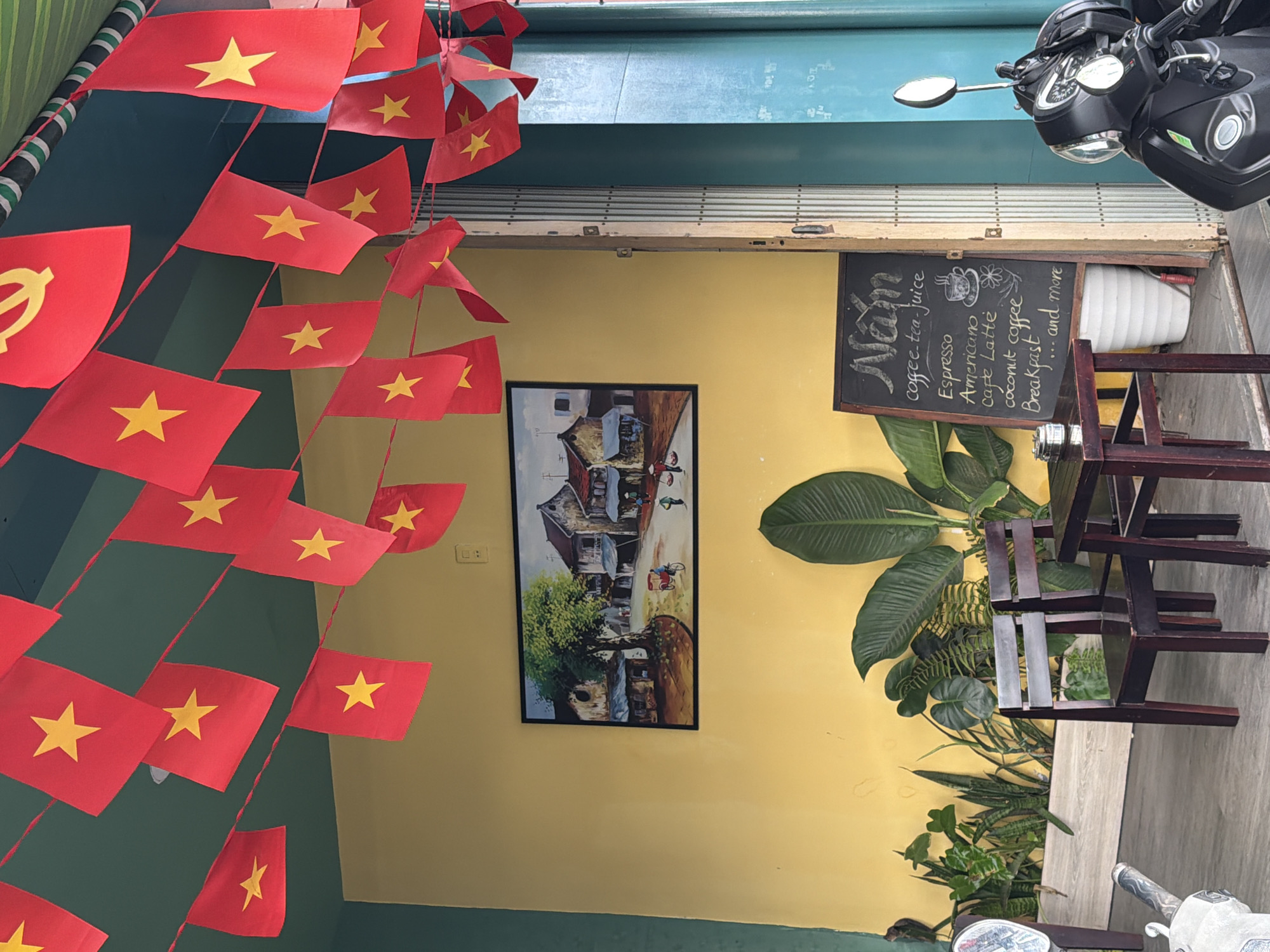 This is a medium shot, eye-level interior view of a cafe in Nha Trang, Vietnam. The space is decorated with red Vietnamese flags with yellow stars strung across the ceiling. The walls are painted yellow and dark green. On the yellow wall, there is a framed painting depicting a street scene with buildings, trees, and people. In the foreground, there are two dark wooden chairs and a small wooden table. To the right of the table, there is a chalkboard menu listing coffee and drink options. Behind the furniture, there are several potted plants, including large-leafed plants and snake plants. To the right of the chalkboard, a metallic roll-up door is partially open, revealing a turquoise wall beyond. A portion of a black motorcycle is visible in the lower right corner of the image. The overall impression is of a relaxed, casual cafe setting.