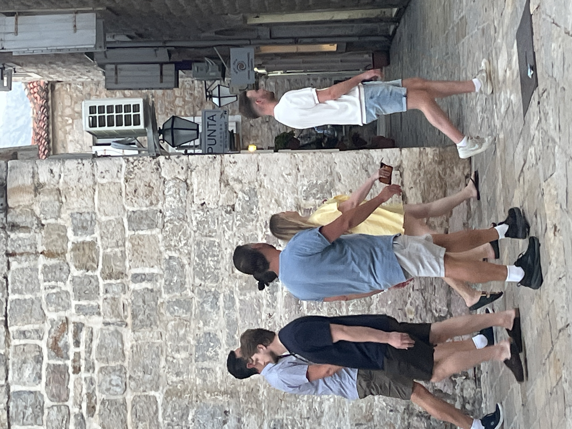 The image depicts a street scene in Budva, Montenegro, characterized by cobblestone pavement and old stone buildings. In the foreground, a group of five individuals are walking. On the left, two men are walking away from the camera. The first man is wearing a light blue short-sleeved shirt and khaki shorts. Behind him, a man in a dark blue t-shirt and dark shorts walks. In the center, a man with his hair tied in a bun is wearing a blue t-shirt and light gray shorts. To his right, a woman in a yellow dress is walking away. Further to the right, a man in a white short-sleeved polo shirt and light blue denim shorts walks away from the camera, looking to his left. Above the group, an air conditioning unit is mounted on the exterior wall of a building, with a black lantern hanging below it. A sign with the word "PUNTA" is visible. The overall impression is a casual stroll through a historic European town.