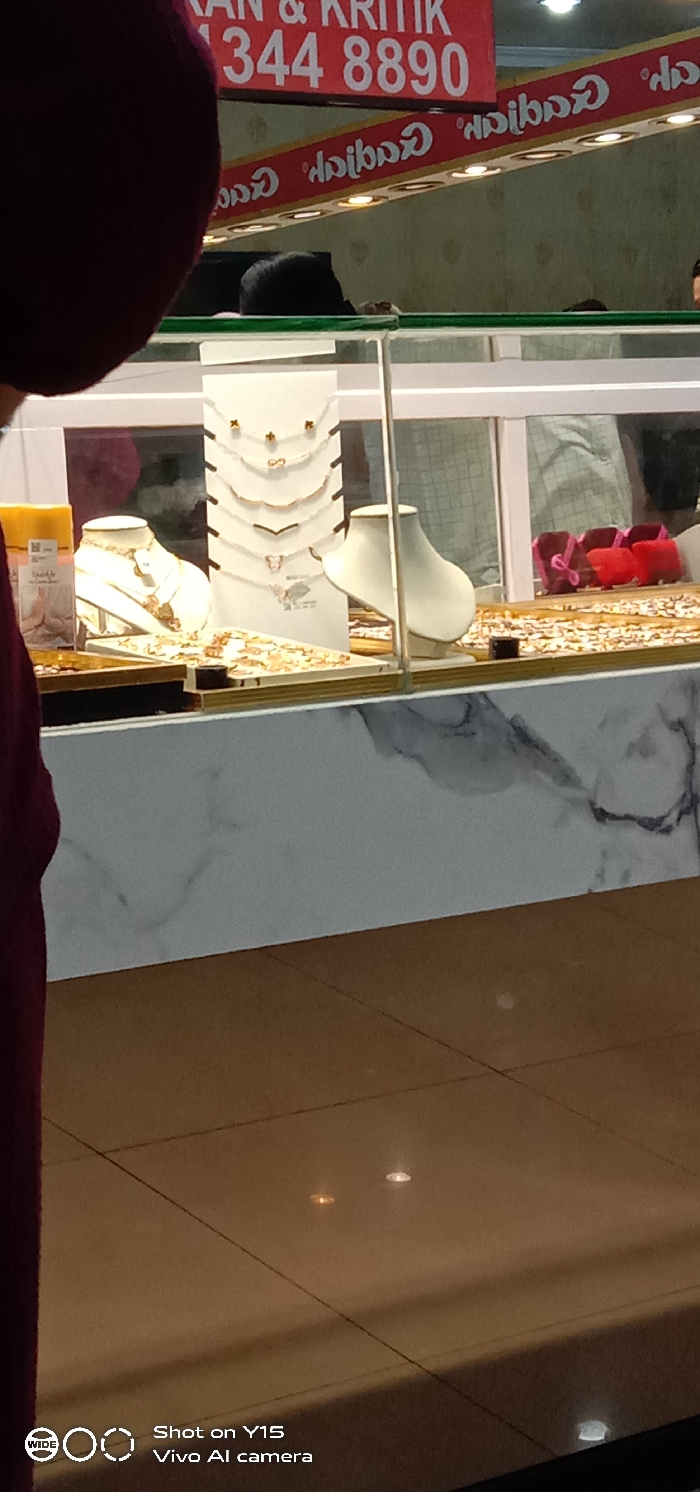 The image displays an interior view of a commercial establishment, identified as "Caribad," in Probolinggo, Indonesia. The foreground shows the left shoulder and head of a person, dressed in dark red or maroon, partially obscuring the view.
The central focus is a glass display case containing various jewelry items. On the left, a white mannequin bust showcases a necklace. Adjacent to it, a white tiered stand holds multiple silver-colored necklaces, some featuring small charms such as a butterfly, a bow, and simple geometric shapes. To the right, another white mannequin bust is visible. The bottom of the display case is filled with numerous small, glittering, gold-toned jewelry pieces, likely rings or earrings. The lower front panel of the display case features a white and grey marble pattern.
Reflected in the glass display case are elements of the surrounding environment, including a person wearing a light-colored, possibly plaid shirt, and several small red objects, likely jewelry boxes or display stands.
In the background, above the display case, two red banners with white text are mounted on a light-colored, patterned wall. The upper banner reads "KAN & KRITIK 344 8890" (Suggestions & Criticisms, followed by a phone number). The lower banner displays the name "Caribad" in reverse, indicating a reflection or brand signage. Small, recessed lights are integrated into the lower banner, illuminating the space.
The floor consists of large, light brown square tiles, exhibiting reflections from overhead light sources. The scene captures a retail environment, implying customer or staff presence within the establishment.