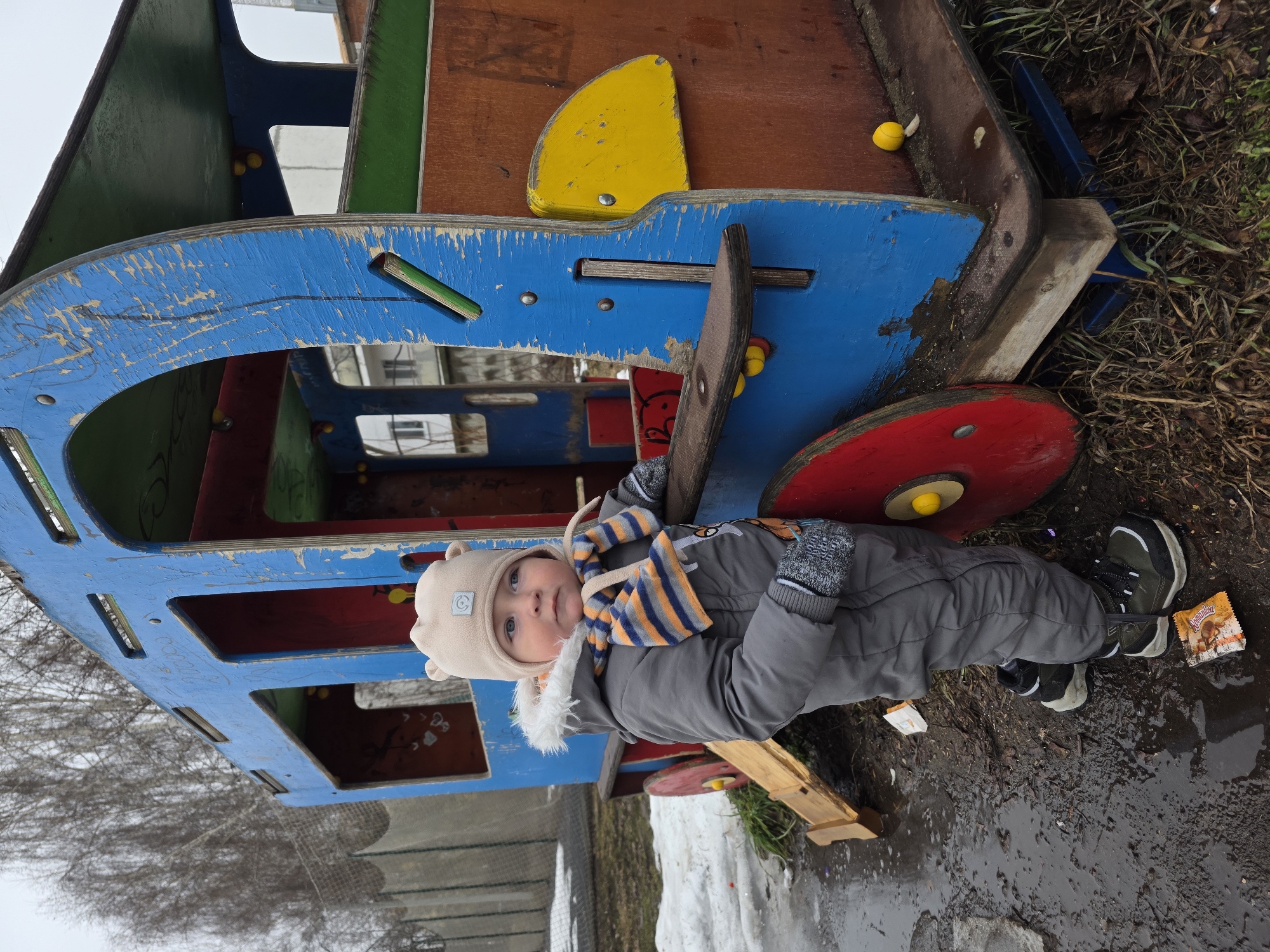 A young child in a gray snowsuit, light brown hat with ears, and an orange and blue striped scarf lies on the ground near a colorful wooden playground structure resembling a train or bus. The structure is painted in vibrant colors like blue, red, yellow, green, and brown. Snow patches are visible on the ground, mixed with wet mud and scattered debris, including discarded wrappers. The background shows bare trees and a glimpse of buildings. The location is Noviy, Russia.