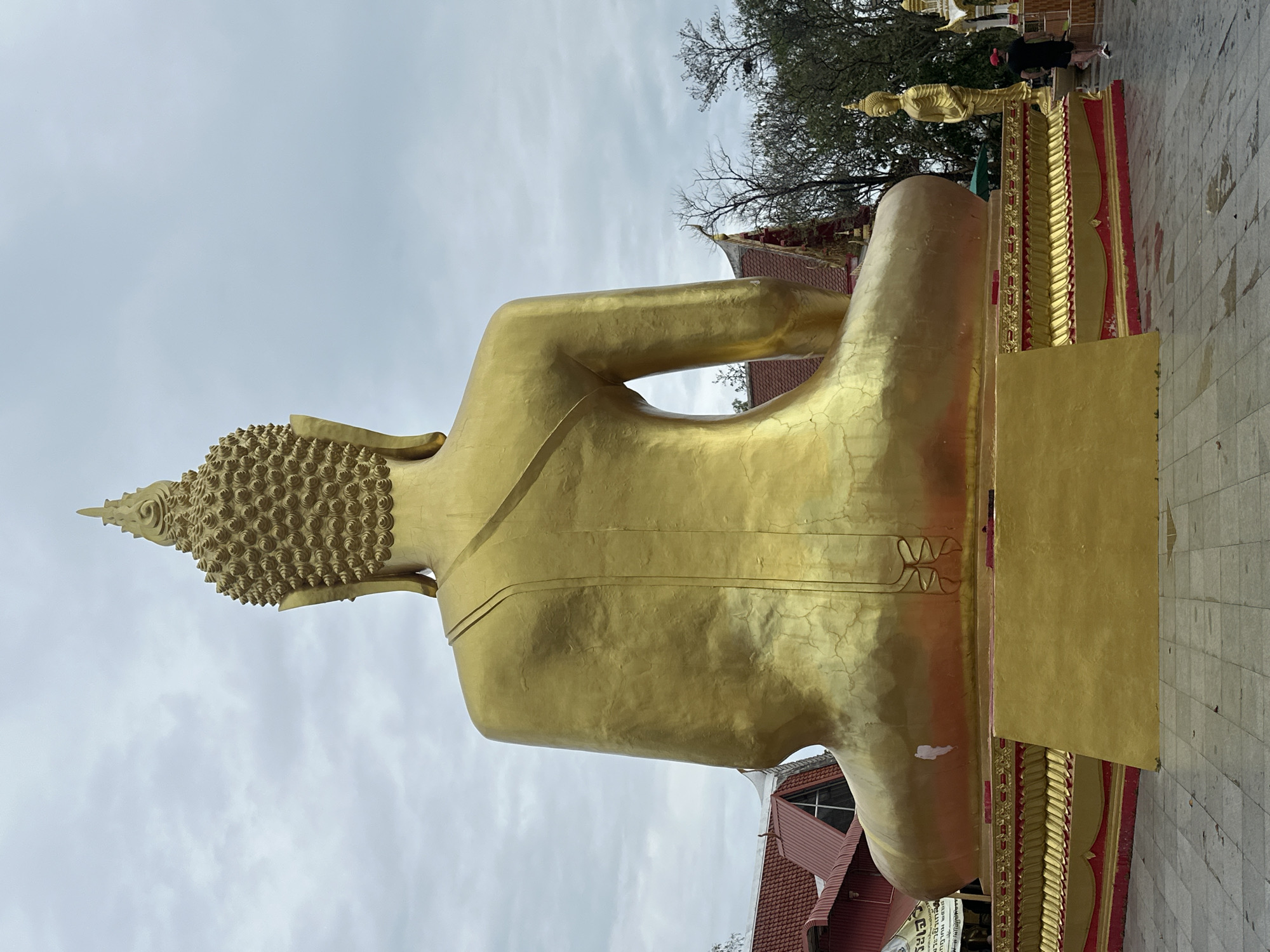 This image shows the back of a large golden Buddha statue located in Pattaya City, Thailand. The statue is positioned outdoors with buildings and trees visible in the background. A person in black clothing is seen walking near smaller statues adjacent to the Buddha. The weather appears overcast.