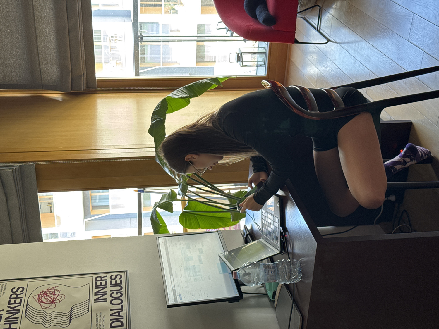 The image captures an indoor scene, likely a home office or study area, illuminated by bright daylight, suggesting it is daytime. The setting is Warsaw, Poland.
The primary subject is a young woman with long, dark hair, wearing a black long-sleeved top and black shorts. She is positioned at a dark wooden desk, leaning forward and appearing deeply focused on a task. Her hands are near an open silver laptop, and an external monitor to its left displays what appears to be text or code. A clear plastic water bottle sits on the desk next to the laptop.
A large, leafy green potted plant, resembling a Bird of Paradise, stands to the woman's left, partially obscuring her face. She is seated on a unique chair with a distinctive, curved backrest made of amber-colored, transparent material. In the background, large windows reveal a view of other urban buildings under a clear sky. Grey curtains frame the windows, and a red armchair is partially visible in the upper right background.
On a white wall to the left, a poster features an illustration of a stylized head with intricate, tangled lines within. The visible text on the poster reads: "MASTERING YOUR THOUGHTS," "HINKERS," "LEARN TO IDENTIFY WHAT YOU CAN AND CAN'T CONTROL," and "INNER DIALOGUES." The overall scene depicts a diligent individual engaged in work or study in a well-lit, organized space.