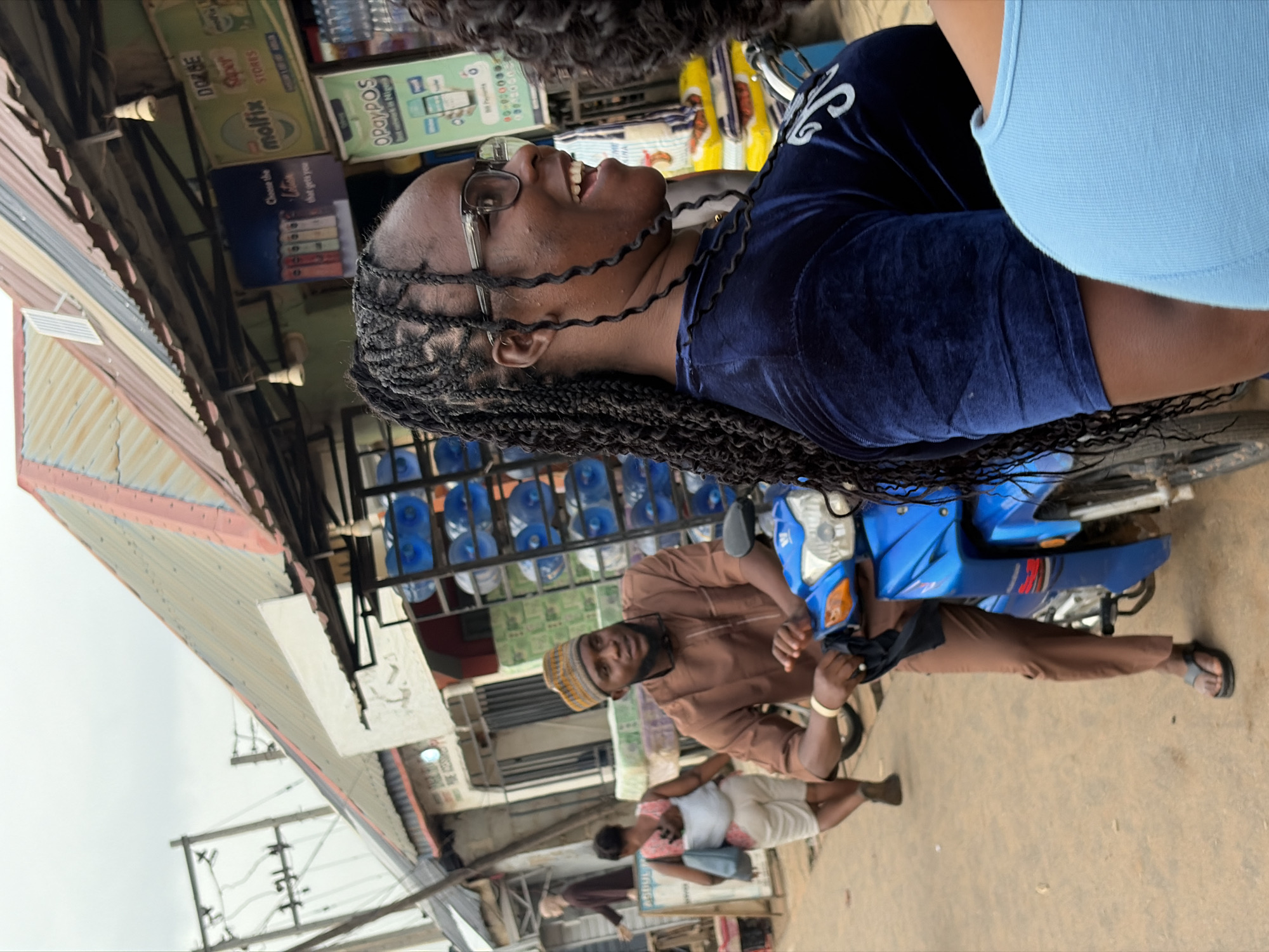 The image captures a street scene in Sabojibi, Nigeria, with a focus on several individuals and their surroundings. In the foreground, a Black woman with braided hair and glasses smiles broadly, wearing a dark blue velvet top. To her left, a Black man wearing a brown traditional outfit and a patterned cap sits on a blue motorcycle, holding a dark cloth. Behind them, another Black woman with a baby wrapped to her back walks away from the camera, wearing a pink and white patterned top and white shorts.
The environment appears to be an outdoor market or roadside vendor area. Shops with corrugated metal roofs line the background, displaying various goods. Prominent elements include stacks of blue water bottles, merchandise on shelves, and advertising posters on the shop fronts. Electrical wires and utility poles are visible in the upper left portion of the image, indicating the presence of infrastructure. The ground is paved with a dirt or gravel-like surface. The overall lighting suggests an overcast day.