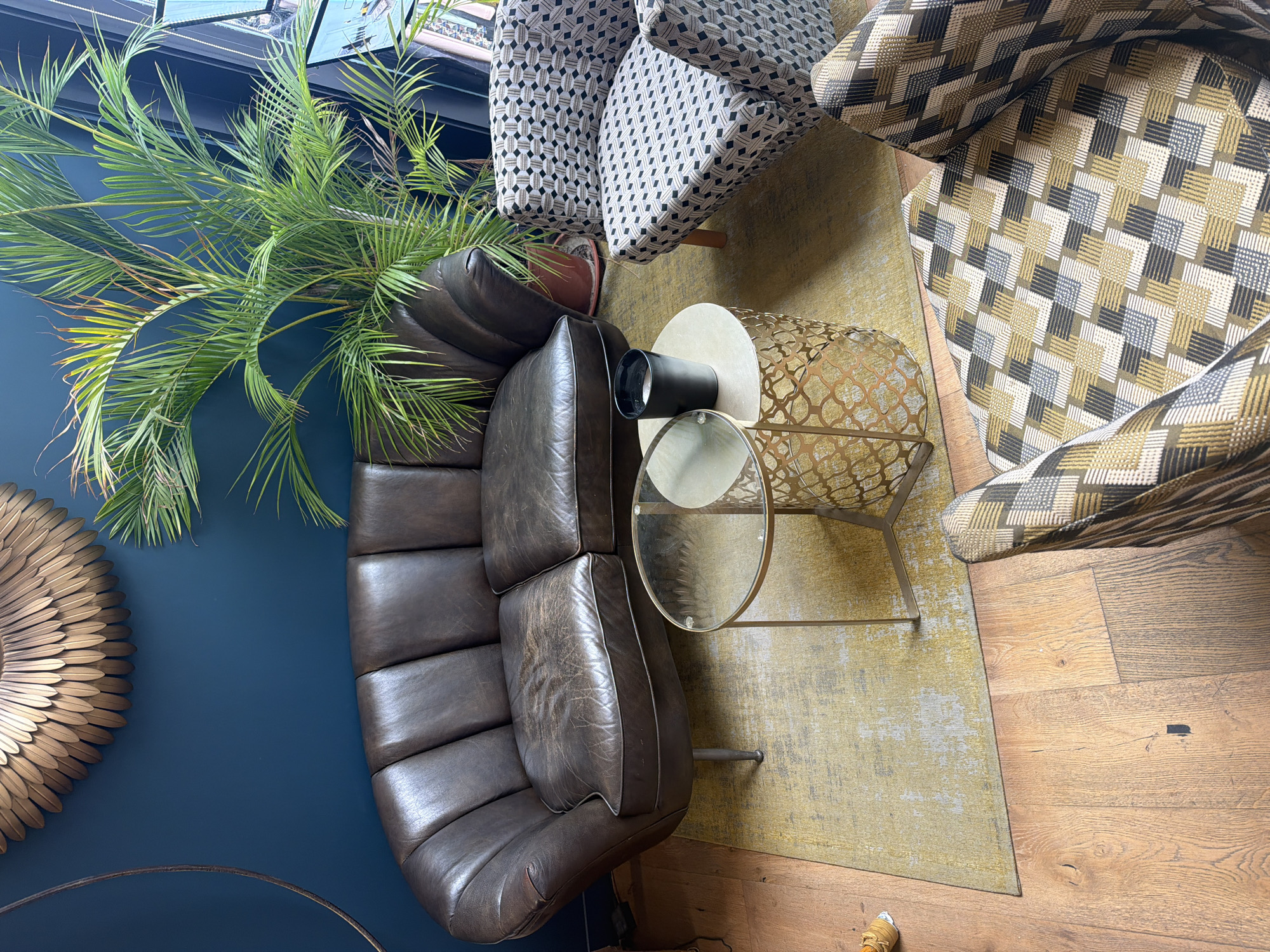 A residential scene, photographed from above, features a dark brown leather couch with multiple segmented cushions positioned against a dark blue wall. Adjacent to the couch is a leafy green plant in a brown pot. A two-tiered end table with a round glass bottom tier and a light-colored top tier holds a cylindrical black object. Two armchairs, one patterned with geometric shapes in black and white, and the other with a similar geometric pattern in shades of yellow, gray, and black, are partially visible at the top of the image. The seating arrangement is set upon a faded yellow rug, which sits on top of a wooden floor. A golden sunburst mirror is affixed to the blue wall to the left of the couch. Given the interior design and the likely origin of the image, it is plausible this scene is set in Paris, France.