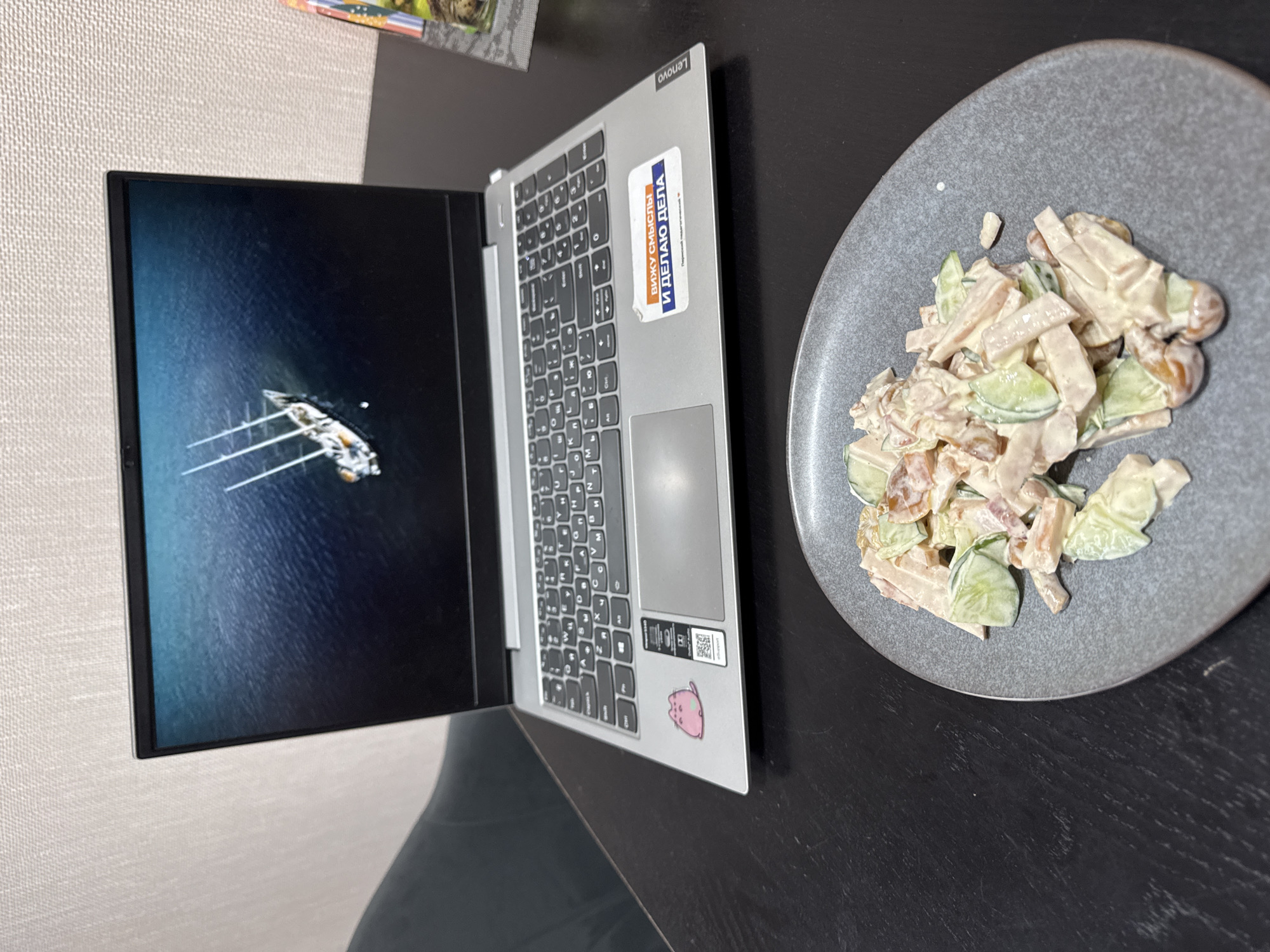 The image shows a laptop and a plate of salad on a dark surface. The laptop is silver and has a Lenovo logo on the side. The screen displays an image of a ship in water. There are stickers on the laptop, including one with Cyrillic text and another with a cartoon character. The plate contains a salad with cucumbers, meat, and a creamy dressing. The location cannot be determined from the image.