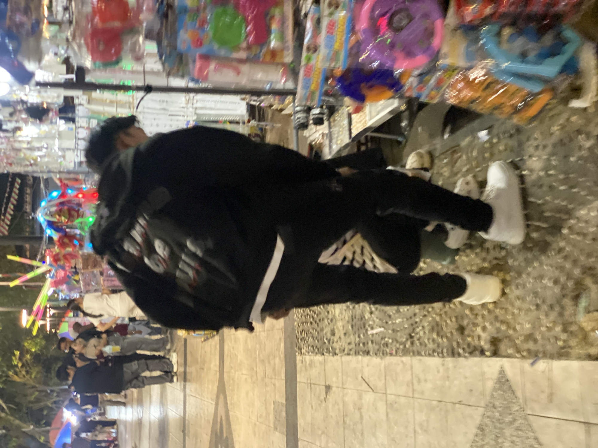 The image captures a street scene in Banjarmasin, Indonesia, likely at night given the artificial lighting and the dimly lit background. In the foreground, a person wearing a black jacket and black pants, along with white sneakers, is partially visible from behind, bent over and interacting with a stall. Their stance suggests they might be examining or purchasing items from the stall, which displays a variety of colorful goods, possibly toys or souvenirs, packaged in plastic. The stall itself appears to be a temporary setup, common in night markets or street vendor areas.
In the mid-ground, a group of people are standing, some looking towards the stall or engaging in conversation. They appear to be shoppers or passersby. The ground is paved with tiles and a section of smaller, rougher stones. The background shows more of the street environment with trees, more stalls, and possibly buildings, illuminated by the lights from the market and streetlights. The overall atmosphere is that of a bustling commercial area at night. The style of the goods displayed on the stall and the presence of street vendors are characteristic of areas like the Siring Tendean or other popular night market locations within Banjarmasin.