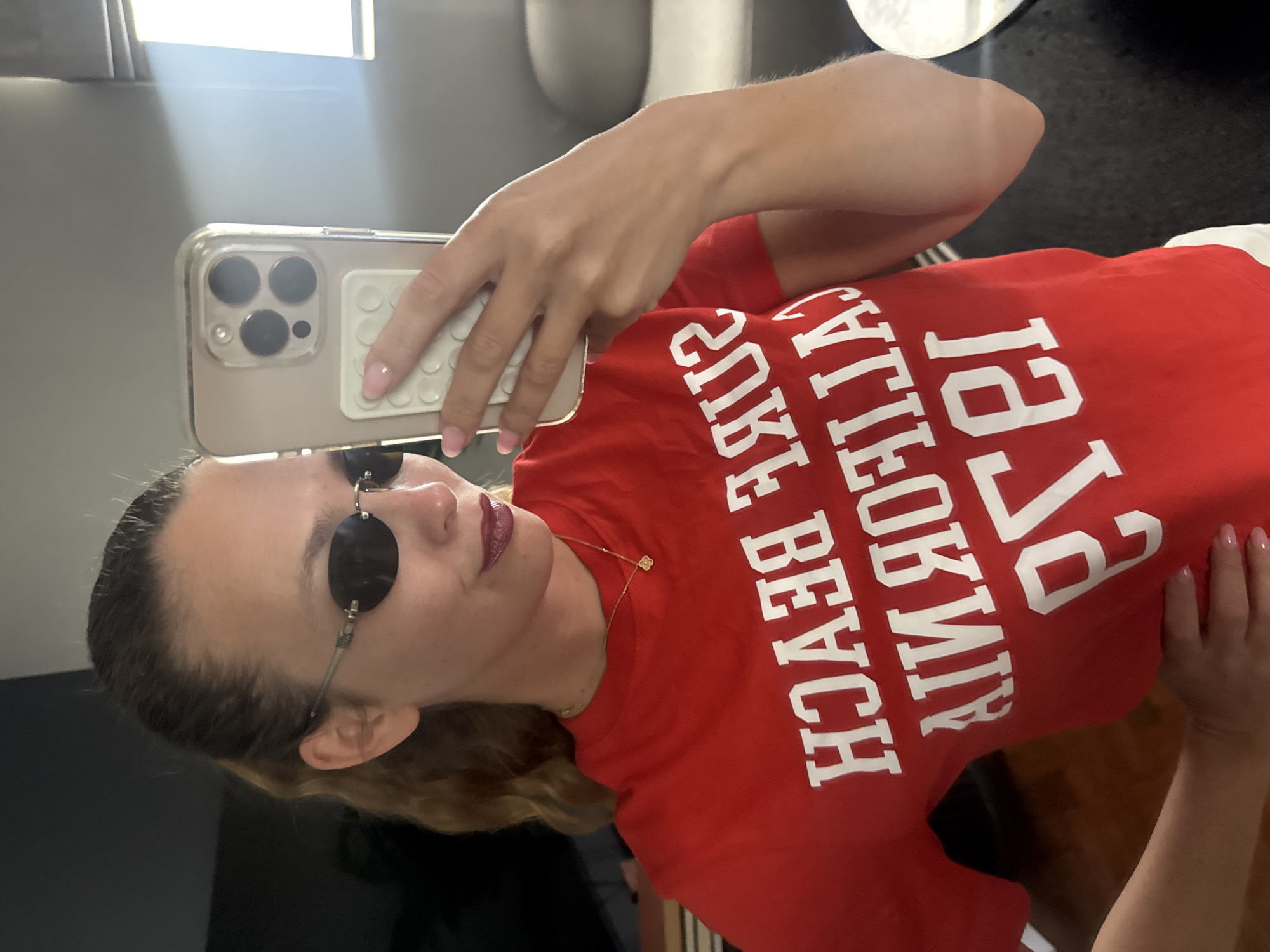 A woman is taking a selfie in a mirror, likely indoors. She is wearing a red t-shirt with white text that reads "1951 CALIFORNIA SURF BEACH". She has sunglasses on and is holding a phone with a clear case and a pop socket on the back. The background is slightly out of focus, but appears to be a room with a neutral-colored wall. There is not enough information to place her in Cape Town, South Africa.