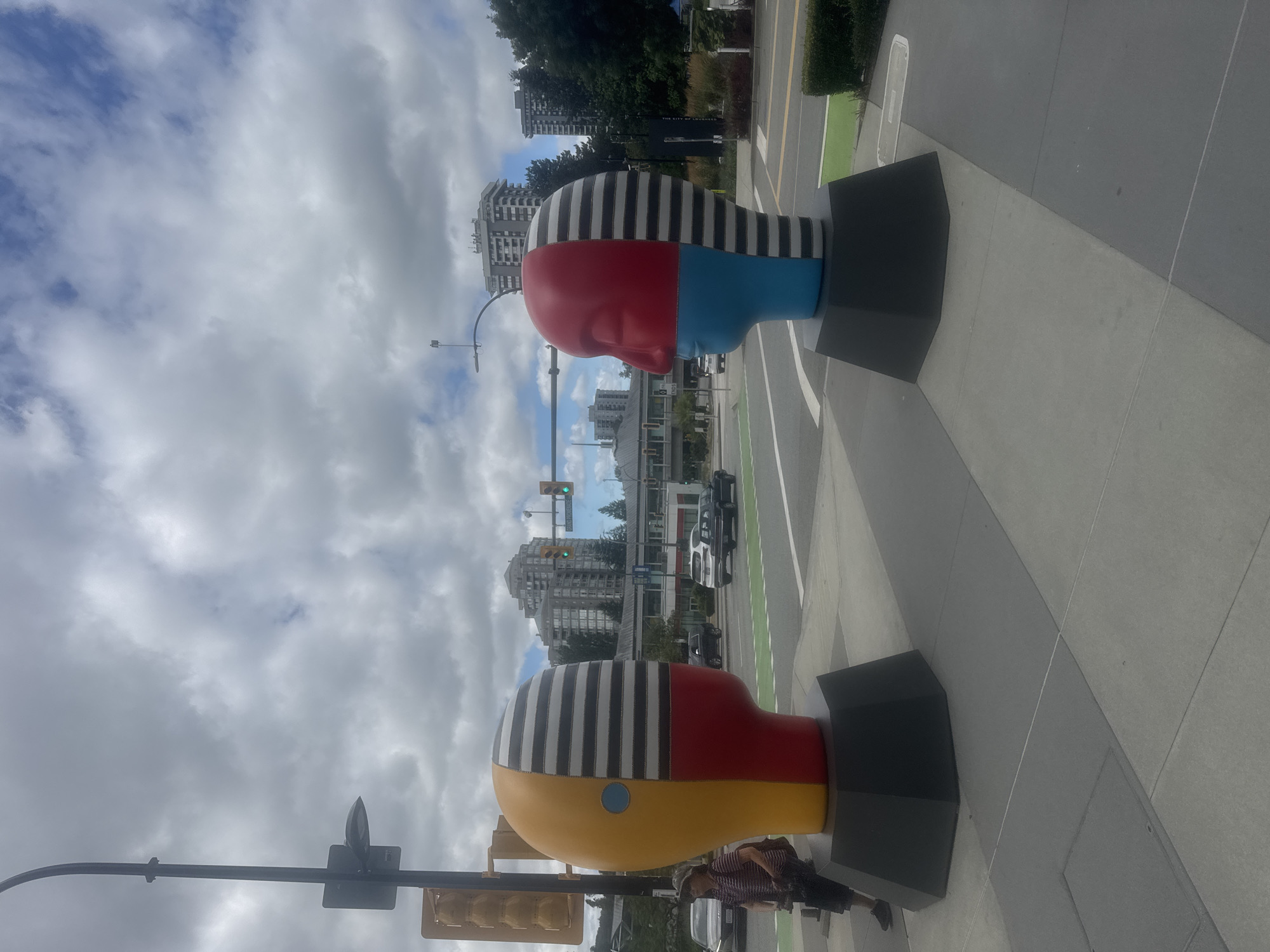 The image shows two large, abstract head sculptures on a sidewalk in Burnaby, Canada. The sculpture on the left is primarily yellow, red, and blue, with black and white stripes on one side of the head. It features a blue circular eye. A person wearing a striped shirt and a hat is standing to the left of this sculpture, looking towards it. The sculpture on the right is red and blue, with black and white stripes on one side of its head. Both sculptures are mounted on dark gray hexagonal bases.
In the background, there are multistory buildings, trees, and traffic lights. Cars are visible on the street, and a pedestrian crossing with green markings is present. The sky is cloudy with patches of blue. The scene appears to be an urban streetscape with public art installations.
