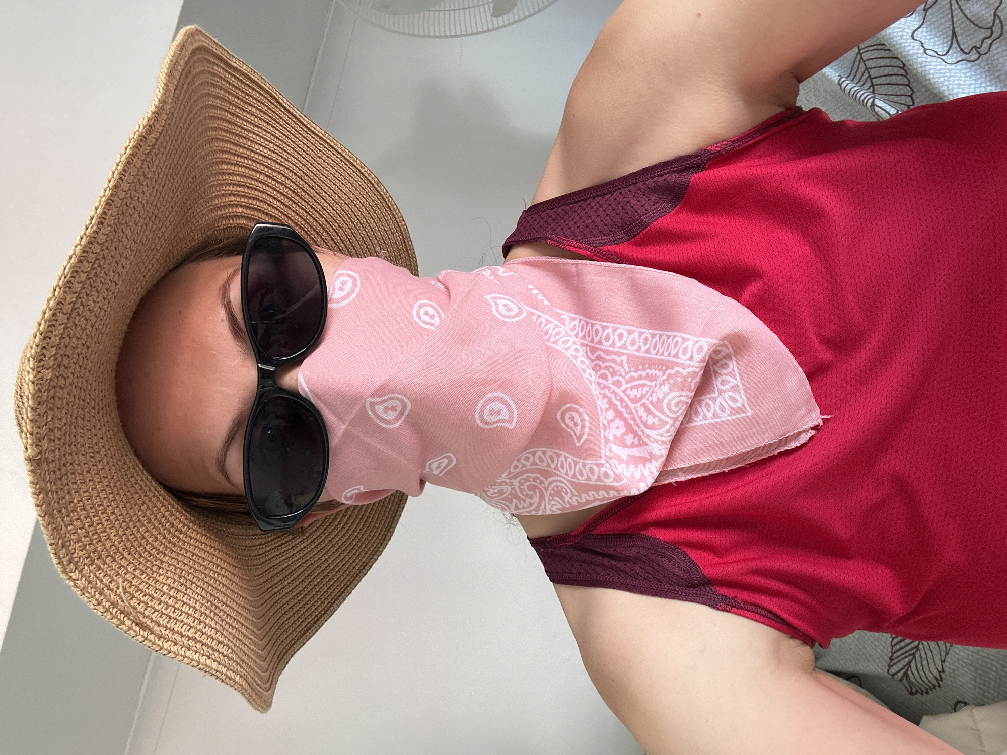 An overhead, slightly angled shot depicts a person in sun-protective attire, likely in a warm climate such as Fethiye, Turkey. The subject wears a wide-brimmed, light brown straw hat and dark sunglasses, which obscure the upper part of their face. A light pink bandana with a white paisley pattern is wrapped around the lower face, functioning as a mask or neckerchief. They are dressed in a short-sleeved, bright red athletic top with maroon detailing around the neck and armholes, suggesting breathable fabric suitable for hot weather.
The background on the left consists of a plain, light-colored wall. On the right, a patterned fabric, possibly a pillow or bedding with grey and white leaf-like designs, indicates an indoor or semi-indoor setting. Bright, even lighting throughout the image suggests it is daytime. The ensemble of hat, sunglasses, and bandana implies readiness for sun exposure or protection from the elements, consistent with a holiday or outdoor activity. No visible text is legible on the subject's attire or in the background.
