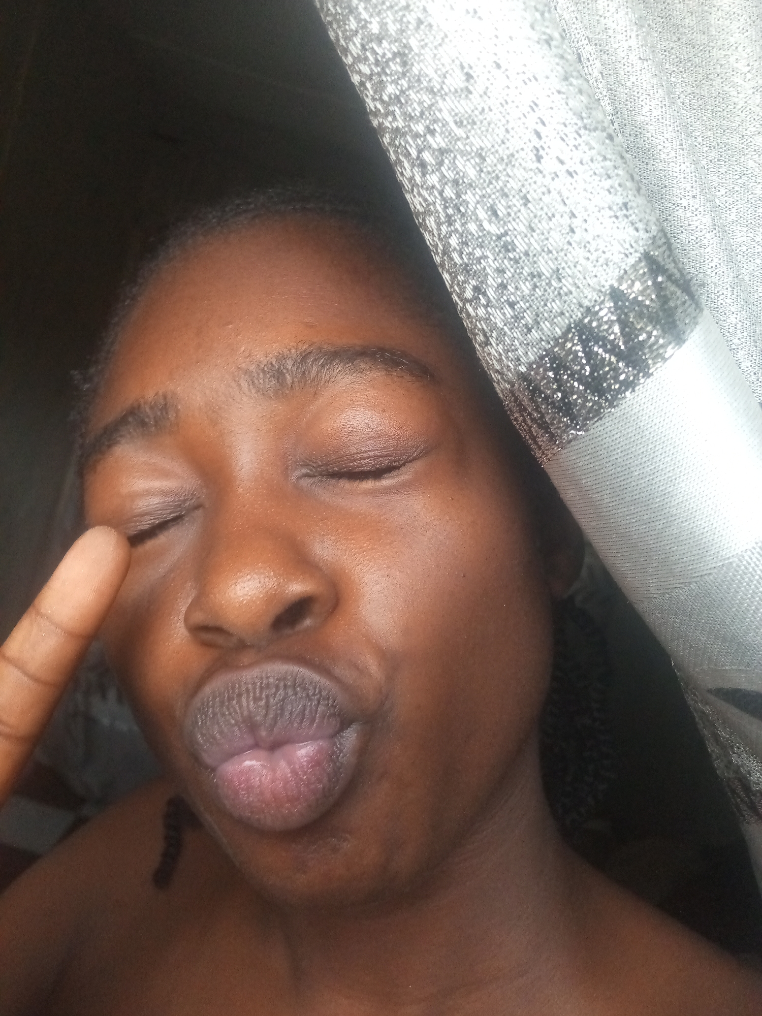 In a close-up, indoor shot, a young Black person with their eyes closed is making a kissing face, with their right index finger touching their eyelid. They have dark hair styled in braids that are visible on their left shoulder, and their lips are slightly parted and colored a light pinkish-purple. The background consists of a dark, out-of-focus area to the left, and a textured, silverish-white curtain with a patterned band on the right. The lighting appears to be soft and diffused, creating subtle highlights on the person's skin.