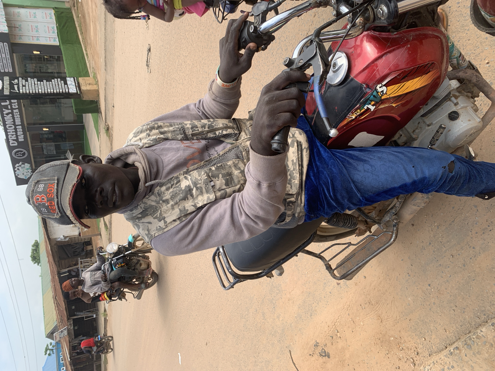 This media file, taken in Shagamu, Nigeria, captures a high-angle, portrait-oriented view of a man riding a motorcycle. The man is wearing a grey hoodie, a camouflage vest, blue jeans with a rip on the knee, and a grey baseball cap with "RED SOX" and numbers visible on it. He is looking directly at the camera with a neutral expression. His hands are on the handlebars, with one hand gripping a lever. A red and yellow sticker is present on the motorcycle's fuel tank, and a blue object, possibly a water bottle, is attached to the bike.
In the background, a young child with braided hair is partially visible at the top of the frame, wearing a pink and white striped top. To the left, a storefront with signage is visible. The text on the signage includes "D'RHONKY_L", "UK/USA UNISEX CLO", "SHOES", and phone numbers. The ground is a dirt or gravel road. Other motorcycles and people are visible further down the road, suggesting a busy street scene. The overall lighting suggests it is daytime, and the weather appears to be overcast. The image is oriented sideways.
